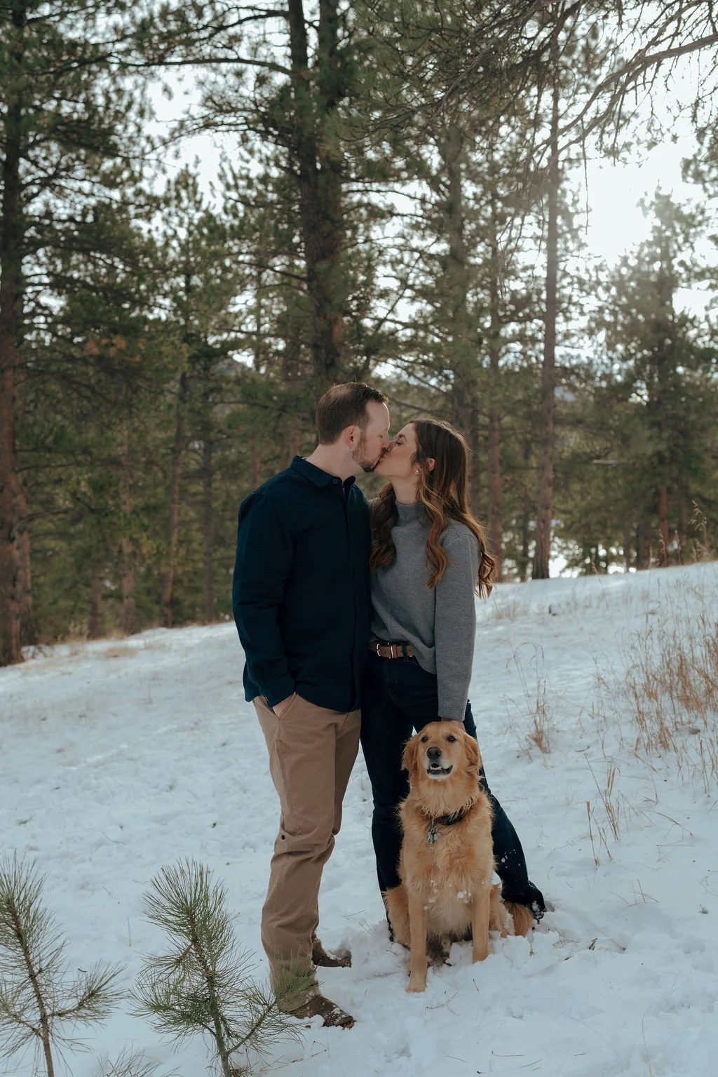 A couple stands in the snow, kissing, with a golden retriever sitting in front of them. Pine trees are visible in the background.