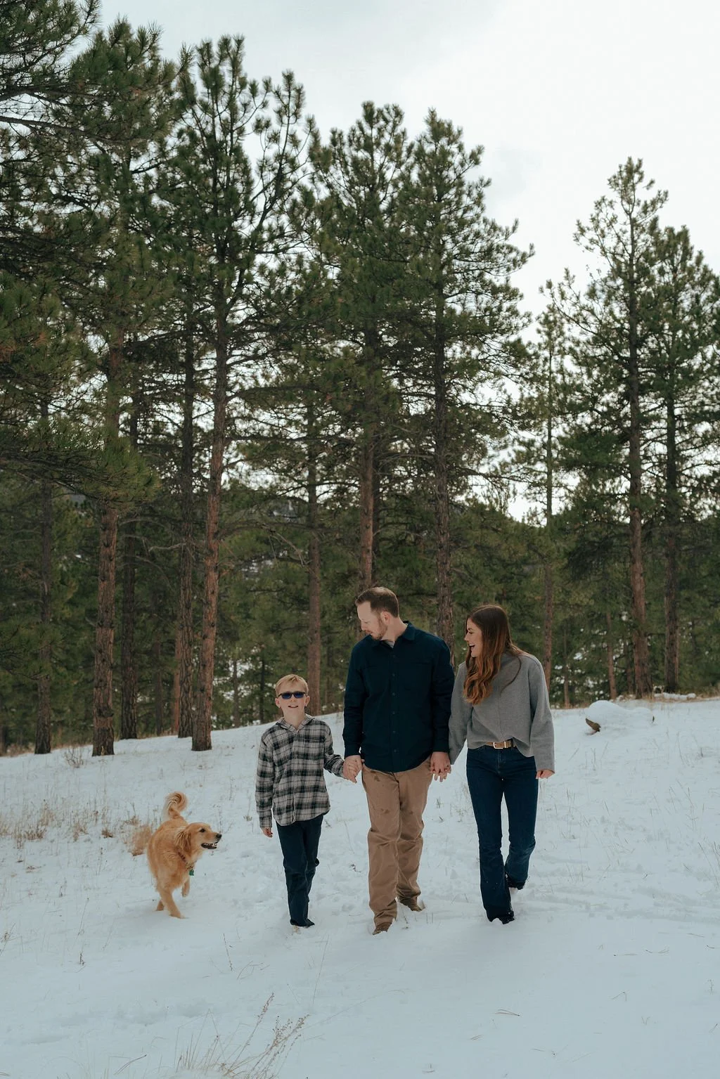 A family of three walks with a dog through a snowy forest, surrounded by tall pine trees for a documentary family photography session