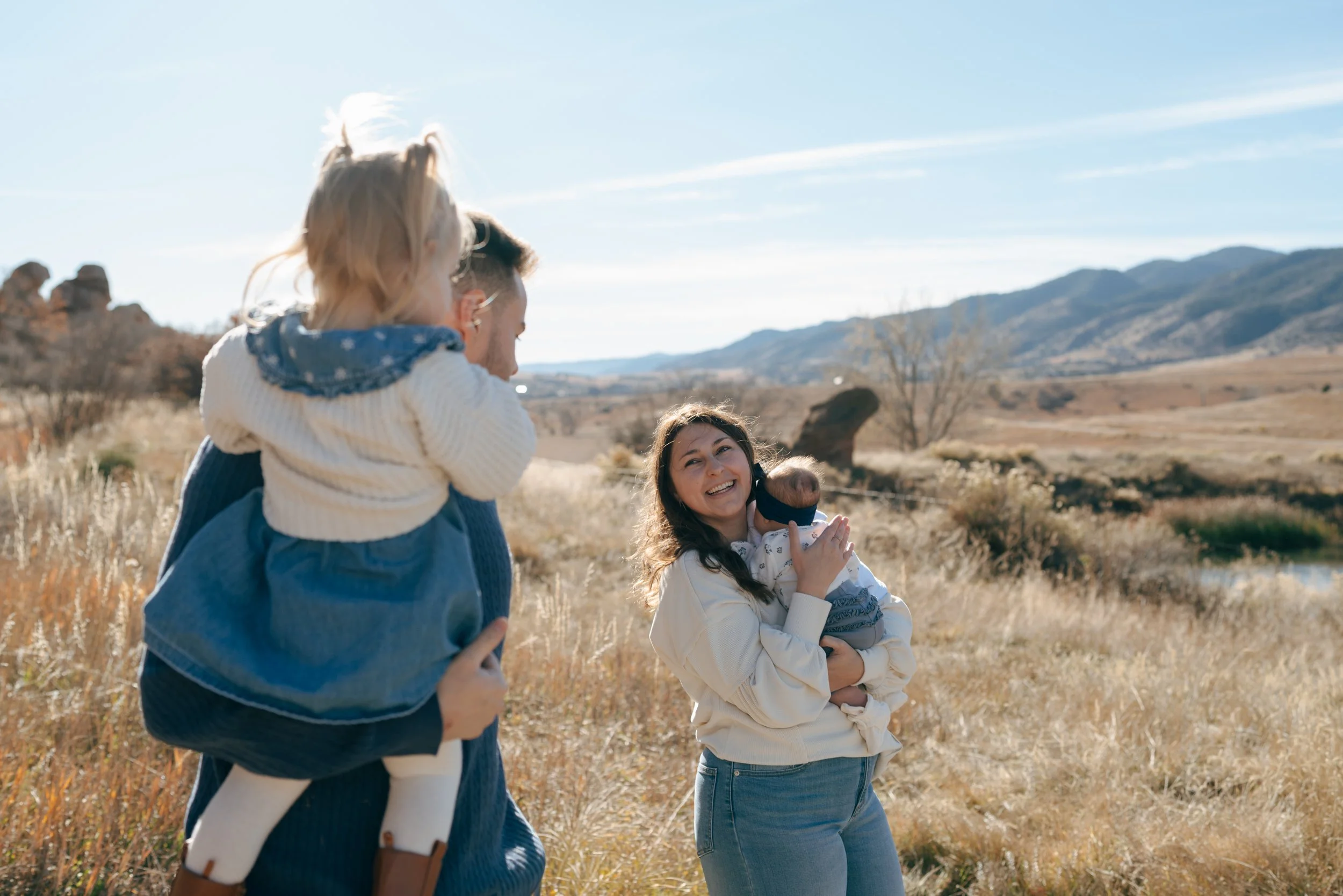 Denver Family Photographer photographs a woman with her baby smiling towards a man with a toddler in his arms with mountains in the background during a fall family session in Colorado