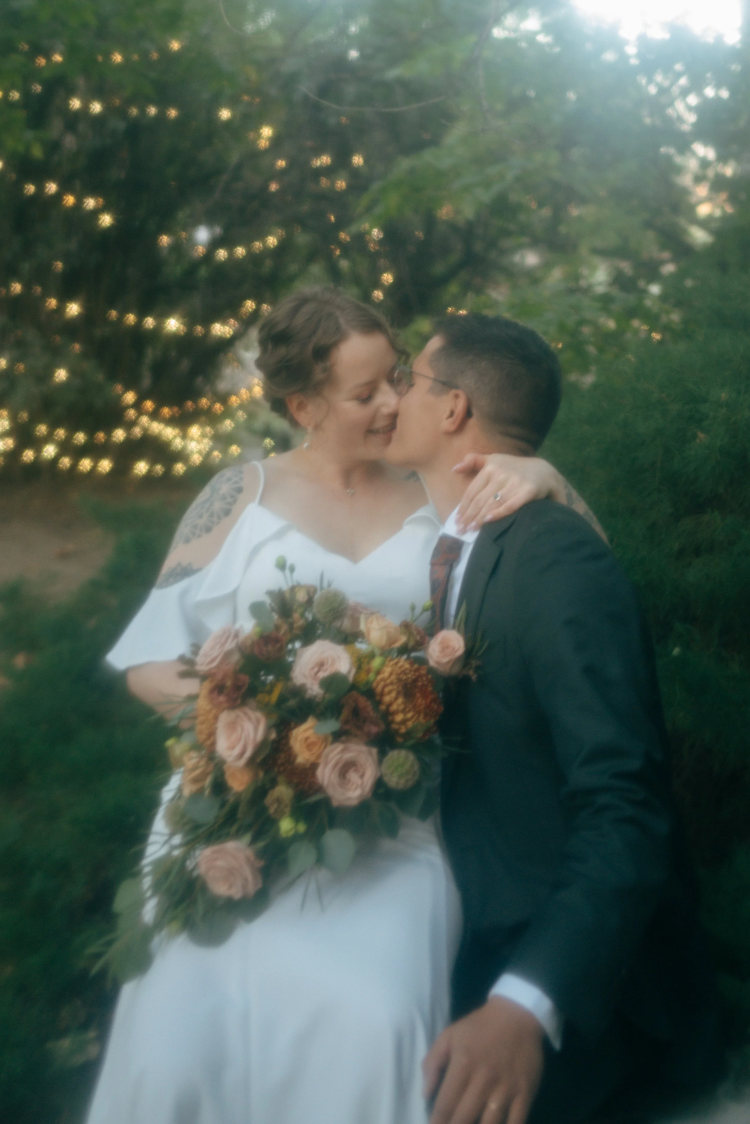 A bride and groom in an outdoor setting with string lights, kissing. The bride is holding a bouquet of flowers with roses and greenery. The groom is in a dark suit, and the bride is wearing a white dress with off-shoulder sleeves.