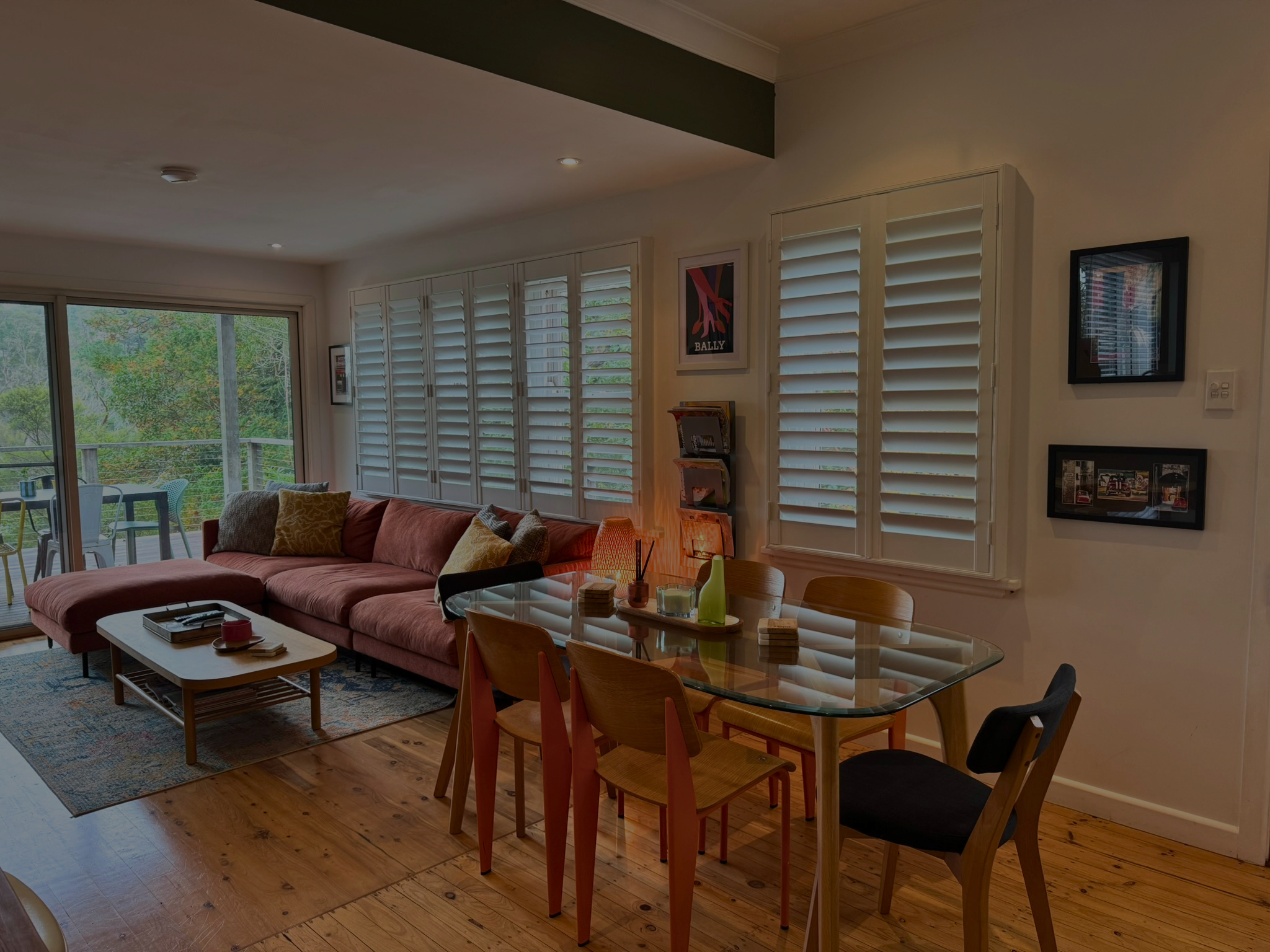 Living room with large orange sofa, wooden coffee table, glass dining table with chairs, sliding glass door leading to outdoor patio, white shutters, magazines on wall, decorative vase, and a floor lamp.
