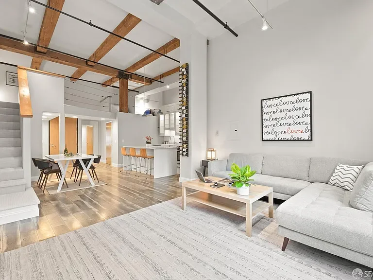 Open-concept living room and kitchen with modern decor, light gray sectional sofa, wooden coffee table with a plant, black and white wall art, and a wooden dining table with black chairs, with exposed wooden beams on the ceiling and hardwood flooring.