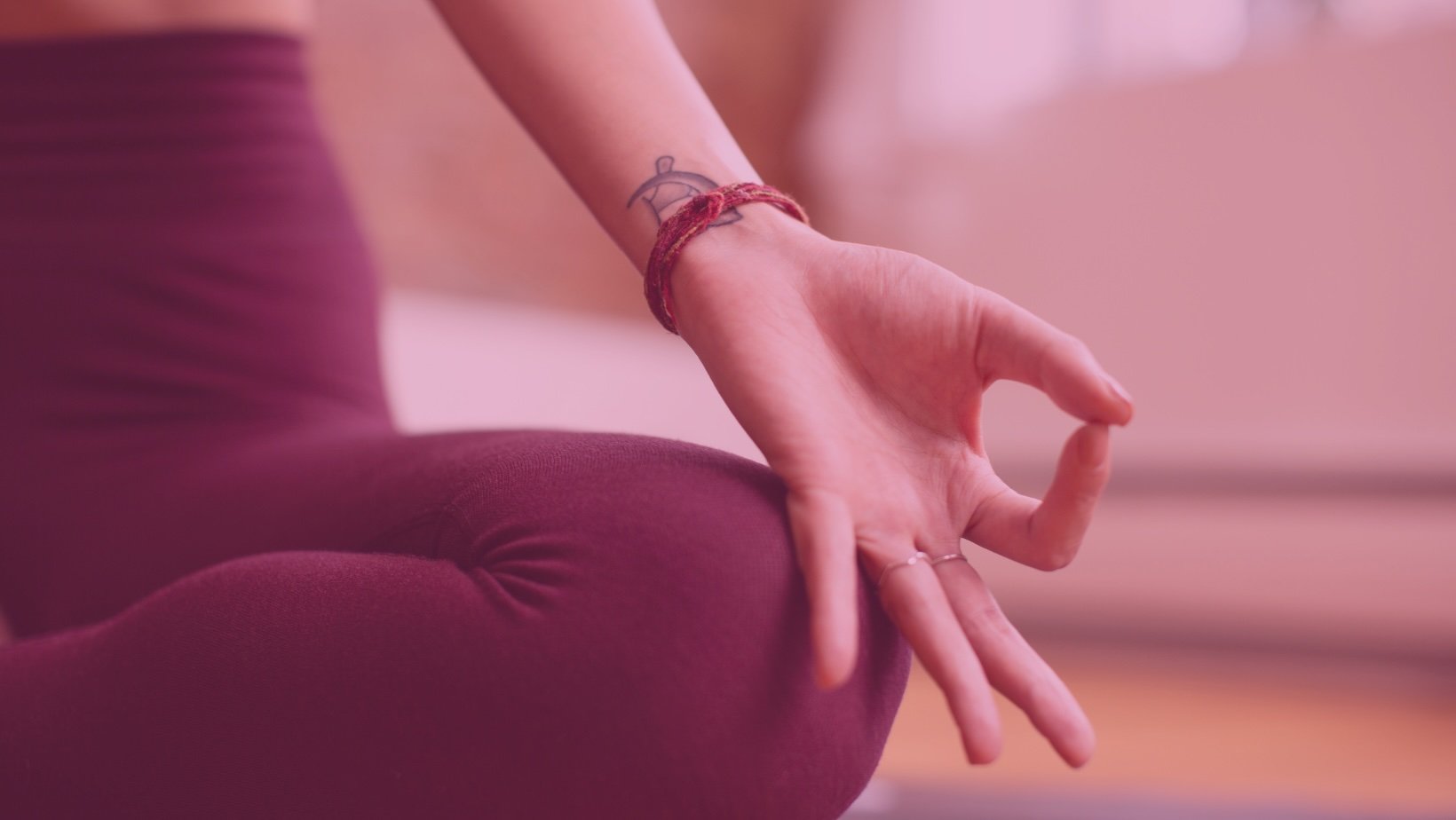 Close-up of a person performing a yoga pose, with focus on their hand and leg, wearing burgundy athletic leggings, with a tattoo on their wrist and a red bracelet.