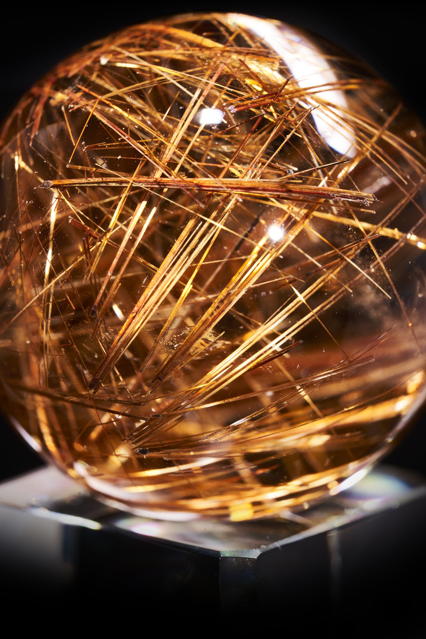 Image Description: A close-up photograph of a high-quality AAA-grade polished Rutilated Sphere Quartz Crystal with red and golden rutile inclusions. The sphere is sitting on a glass stand.