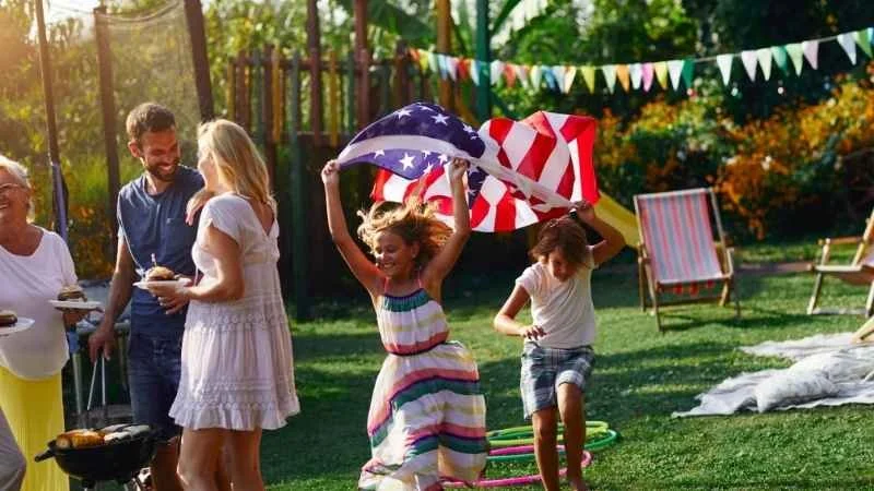 A garden party with people chatting and two young children carrying the American flag