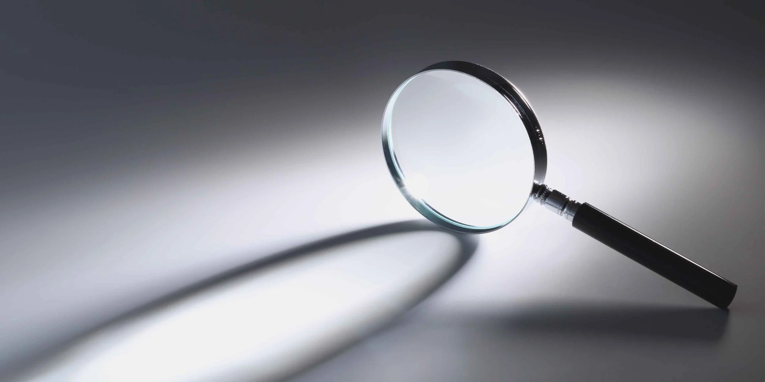 Close-up of a magnifying glass illuminated by a bright light, casting a long shadow against a dark grey background.