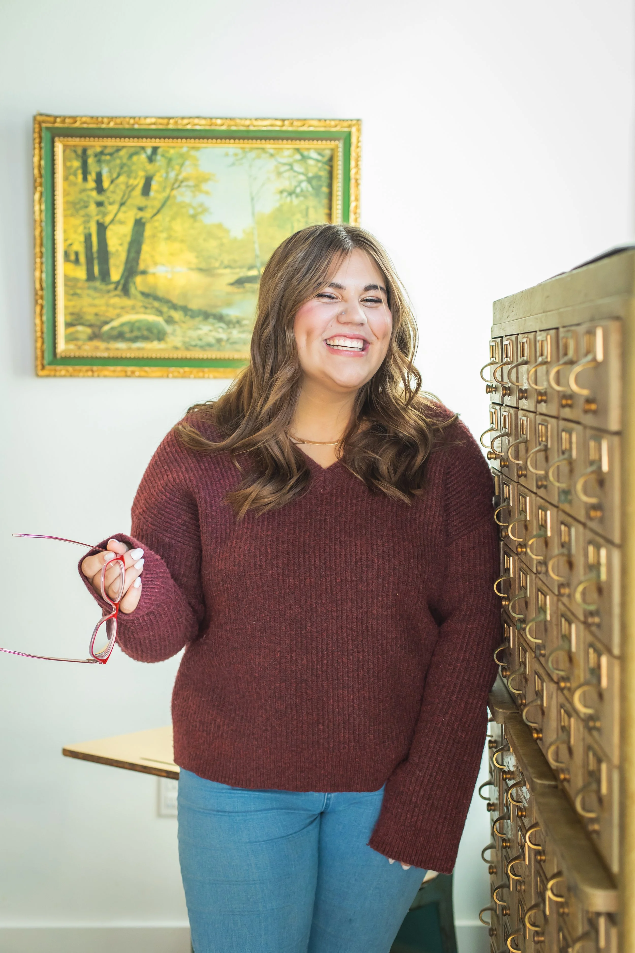 A young woman with long, wavy brown hair is smiling and holding glasses. She is wearing a burgundy sweater and light blue jeans. There is a painting of a forest scene hanging on the white wall behind her.
