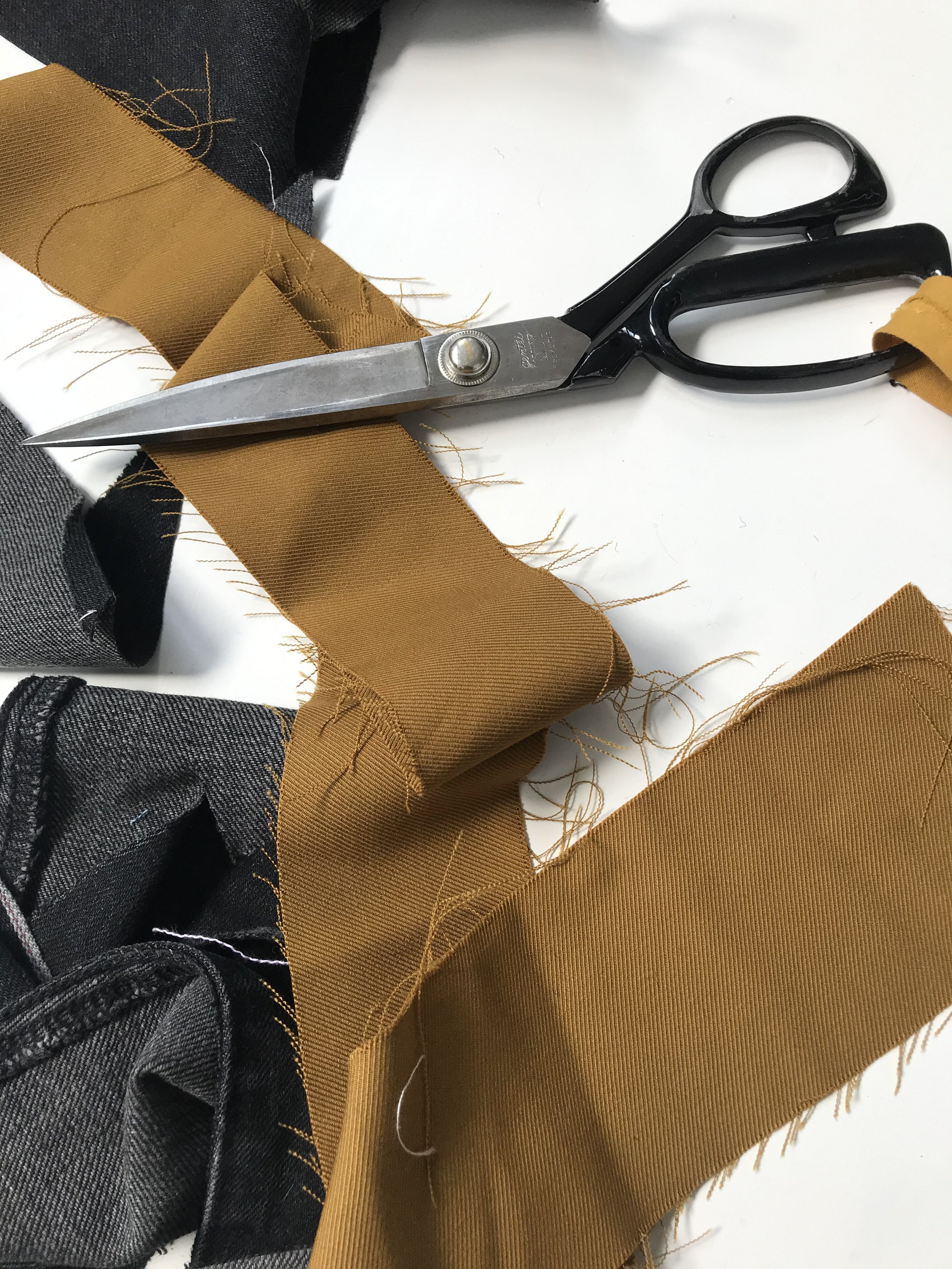 Une paire de ciseaux posée sur des tissus en coton de différentes couleurs, dont noir, gris, et moutarde, avec des bords effilochés.