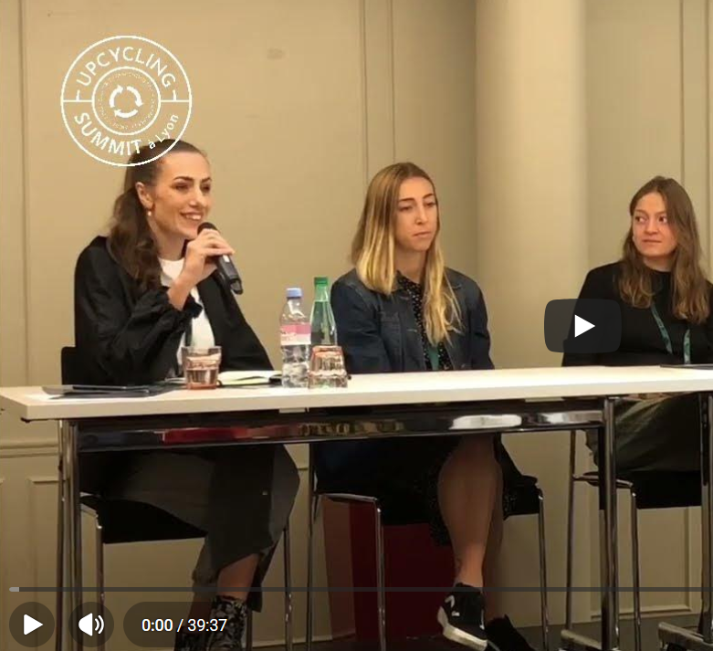 Trois intervenantes assises à une table lors d'une table ronde à Lyon. Lucie Mestre parle dans un micro, les deux autres écoutent. Il y a des bouteilles d'eau et des verres sur la table. Logo « Upcycling Summit » en haut à gauche.