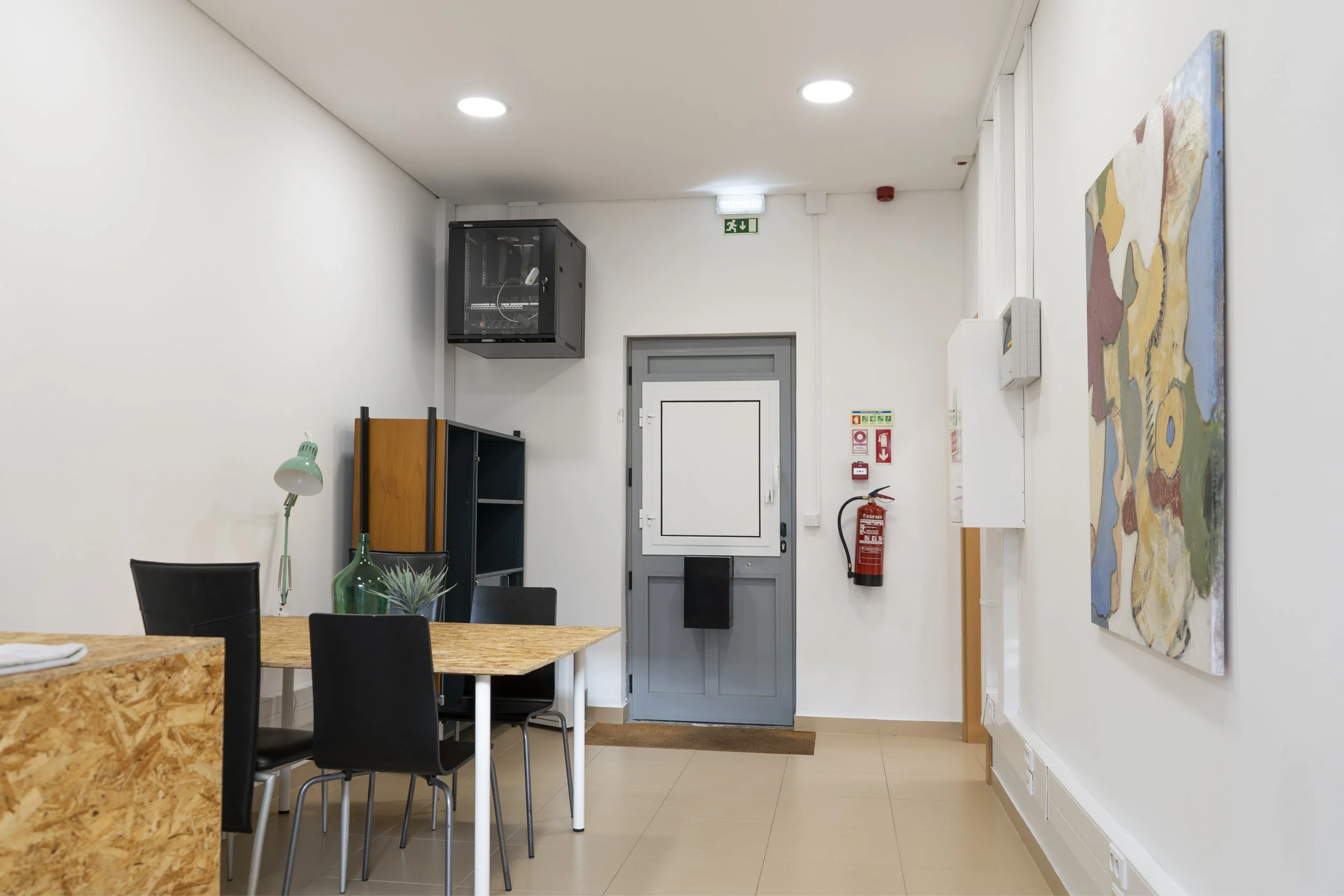 Interior de uma sala com paredes brancas, uma mesa de madeira com cadeiras pretas, um candeeiro de secretária verde, garrafas decorativas, uma pintura abstrata colorida na parede, uma porta cinzenta com uma pequena janela, equipamento de combate a incêndios e um sinal de saída de emergência no teto