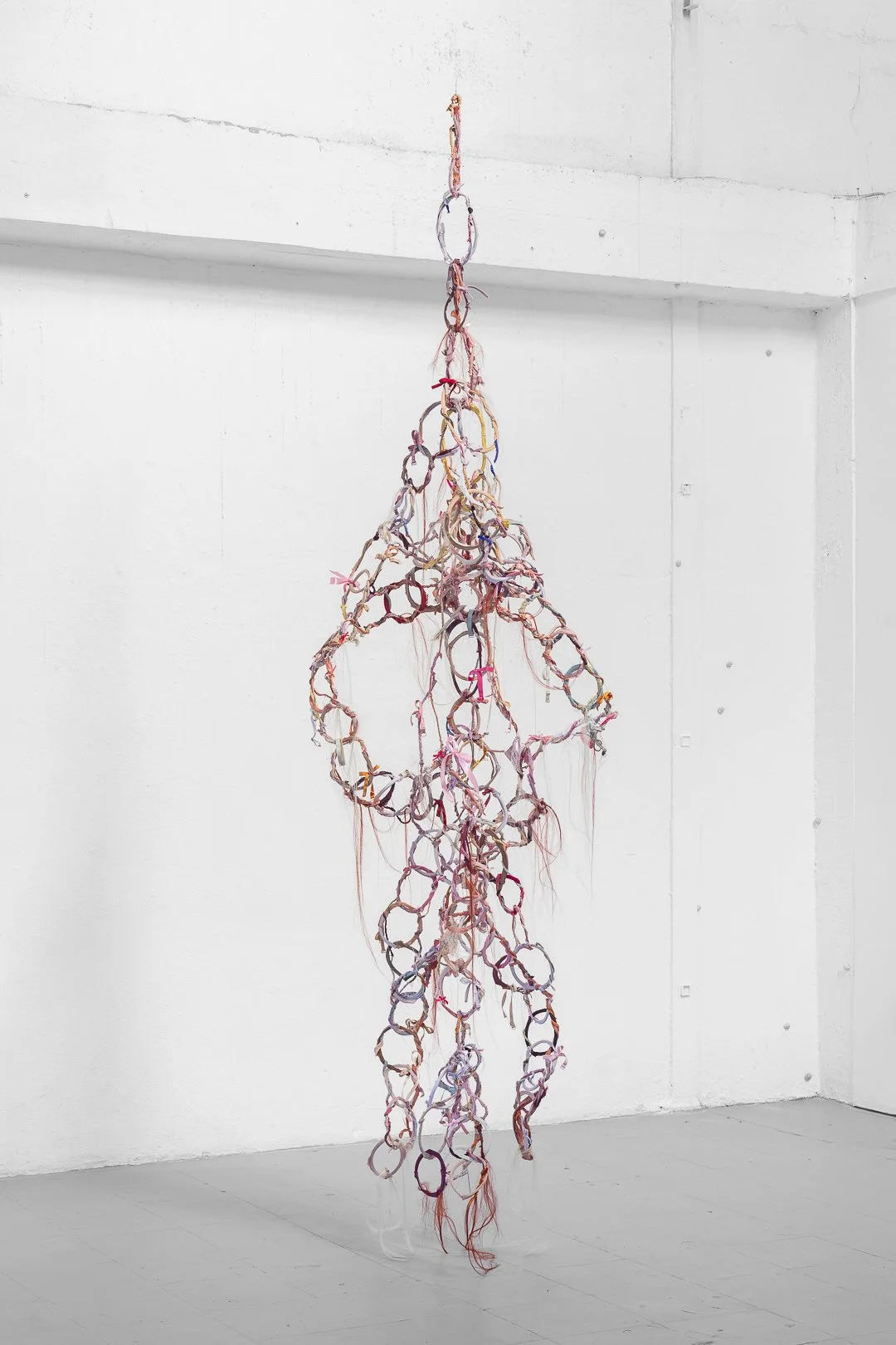 Janna Yotte, Ce qui soigne, soutient et bouche le drain. [What heals, supports and clogs the drain.] Bra wire, used textile, thread, artificial hair, wire, 330 x 89 x 53 cm, 2026.