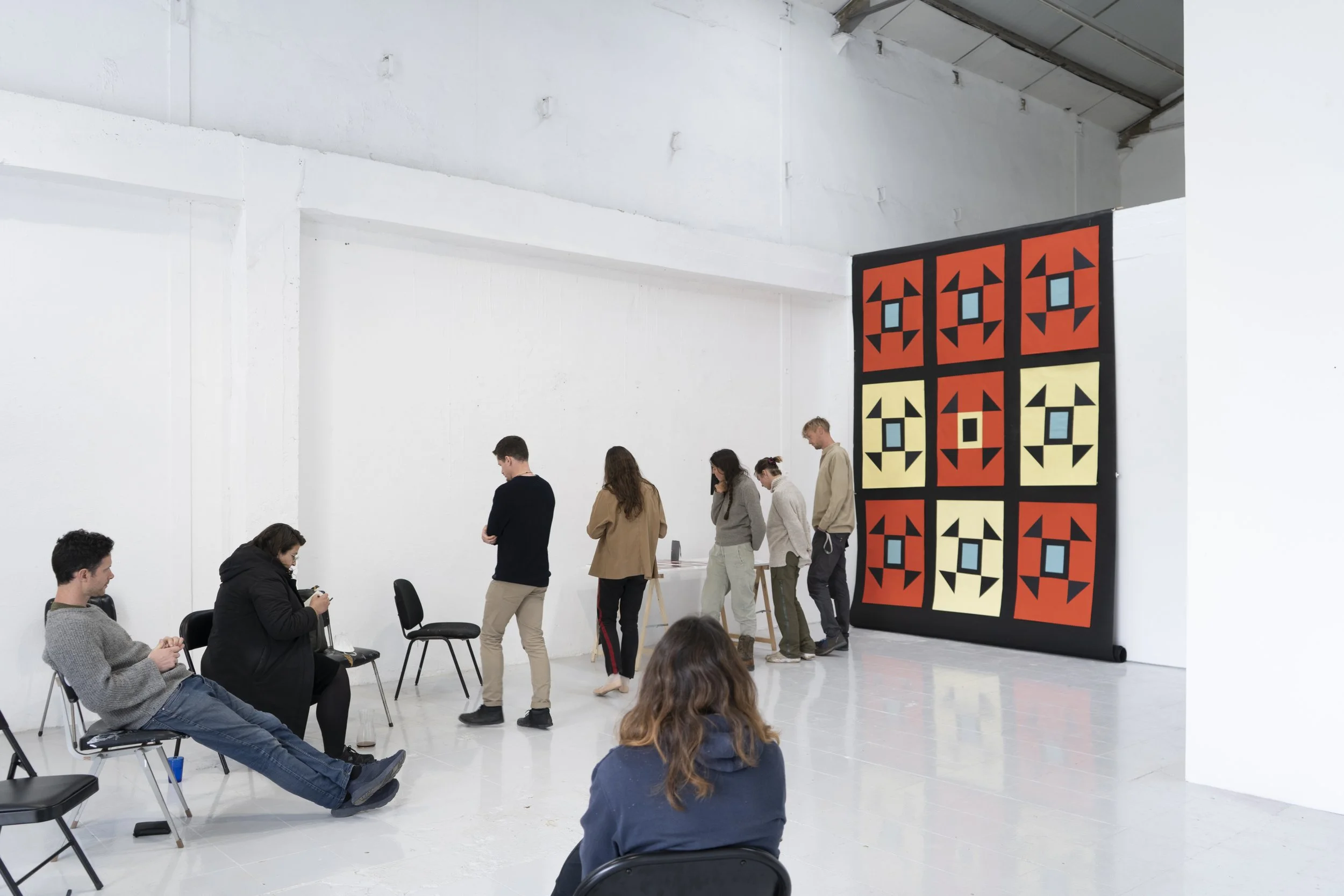 Pessoas sentadas e de pé numa galeria com uma grande exposição de colchas na parede.