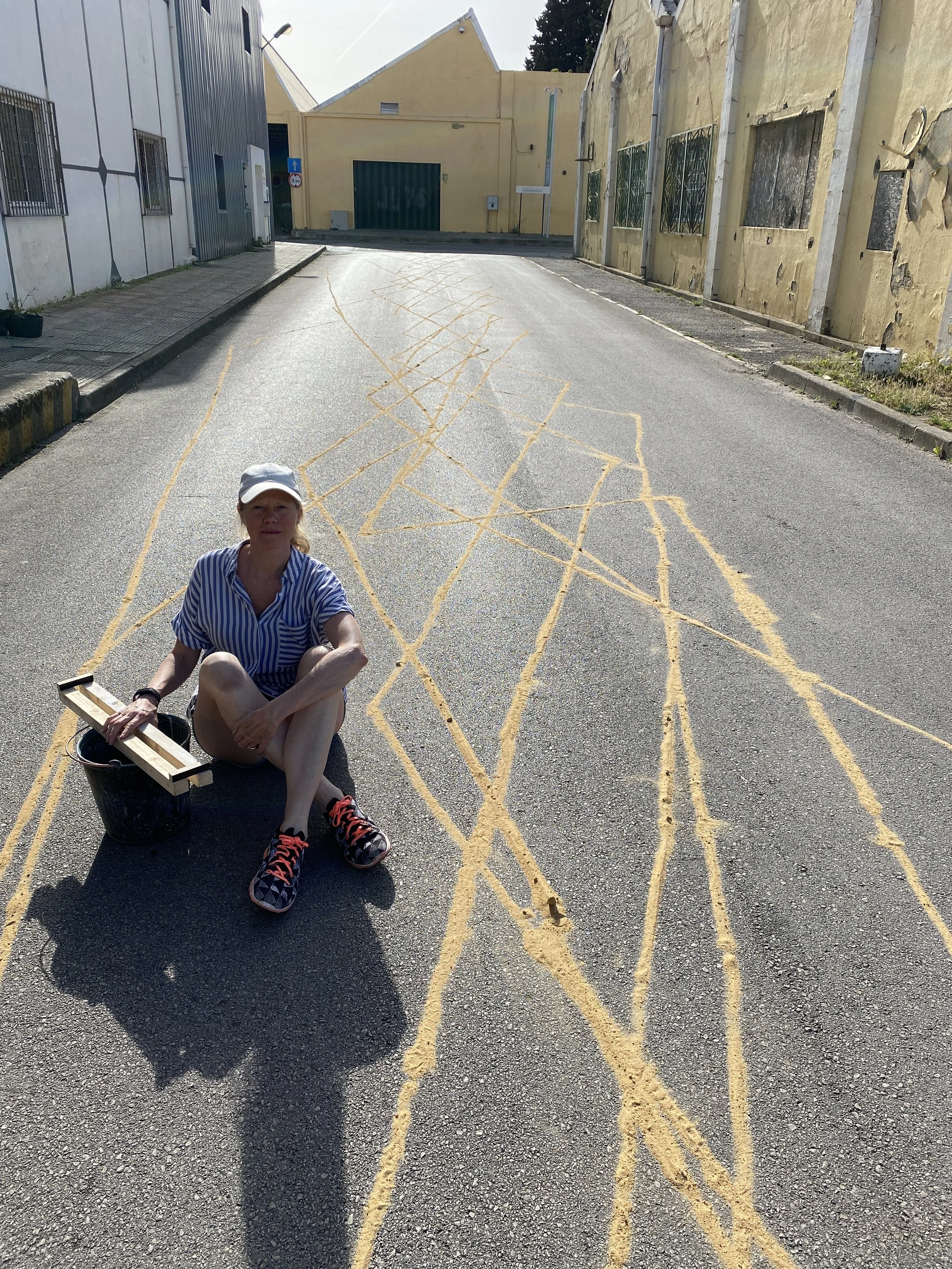 Alice Sheppard-Fidler, sitting on the floor next to a sand sculpture on a gray road.