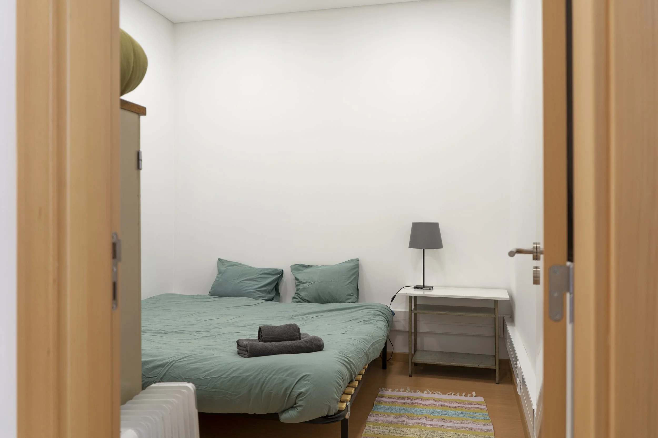 Quarto minimalista com uma cama verde, duas almofadas a condizer, toalhas cinzentas dobradas em cima da cama, uma secretária branca com um candeeiro cinzento, um pequeno tapete multicolorido e chão de madeira clara, visto da entrada.