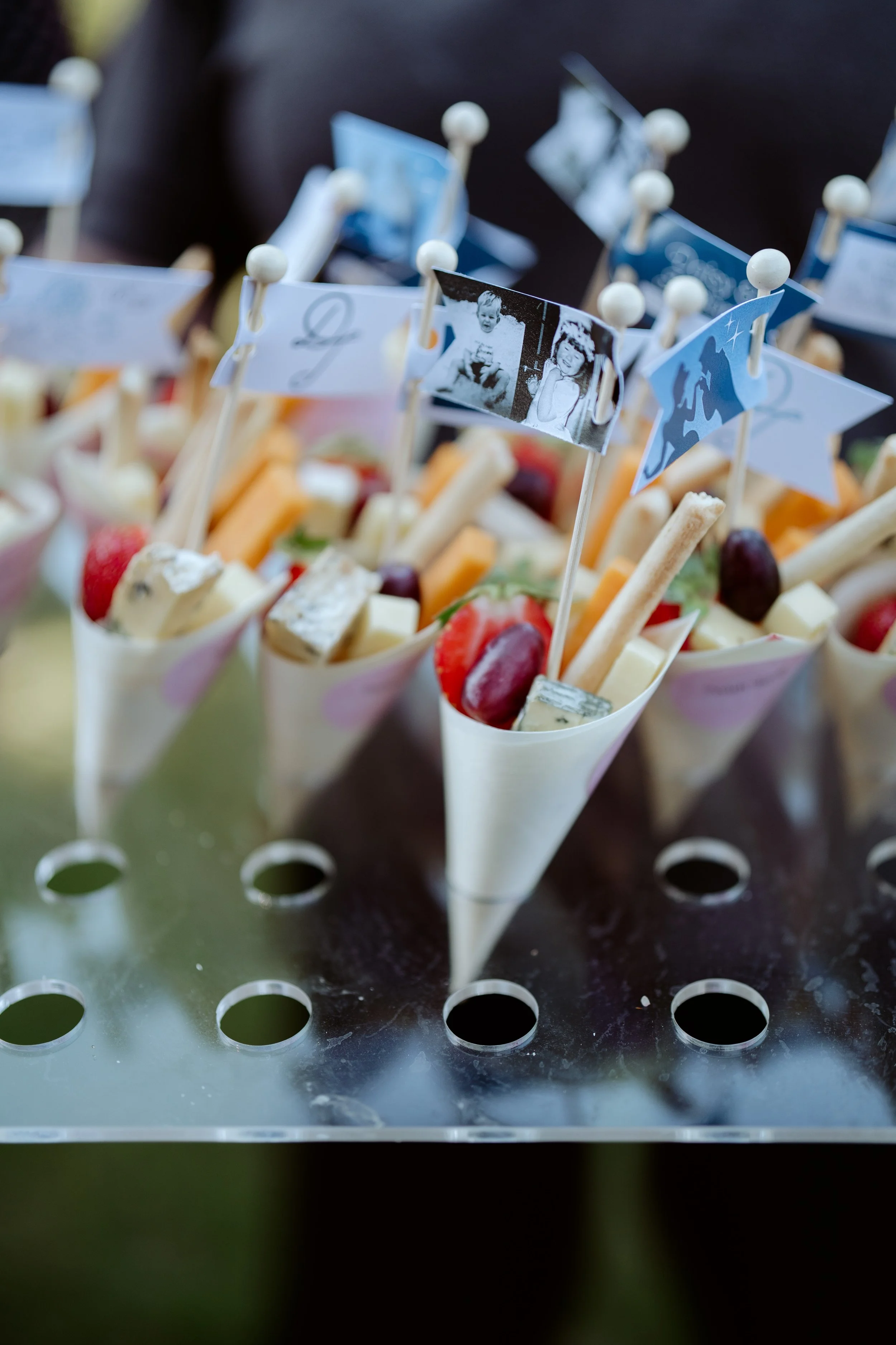 Multiple small white paper cones filled with cheese, fruit, and crackers, each with a small flag with black-and-white vintage photos and blue graphics, placed on a black perforated metal surface.