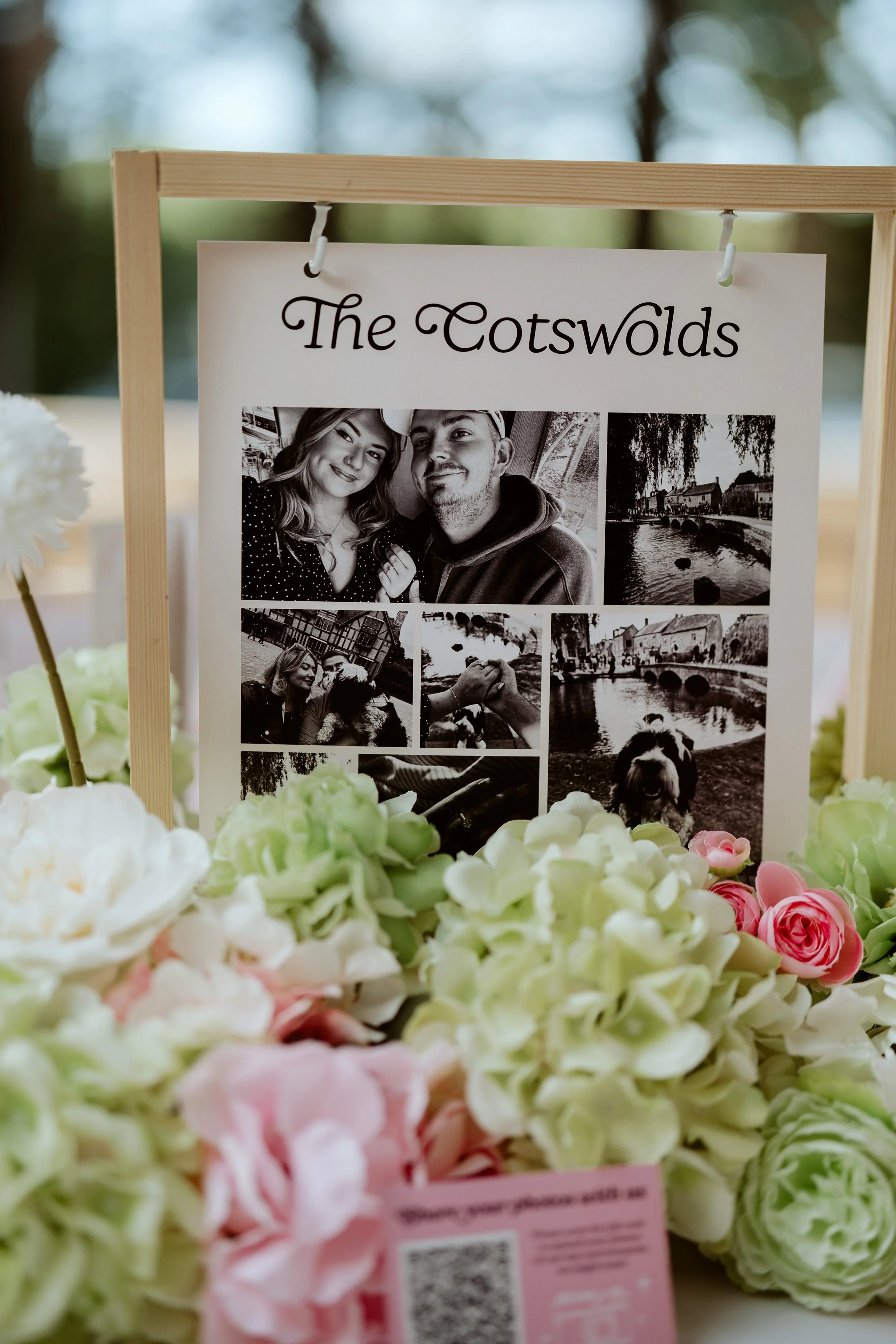 A framed display with black and white photos and the text "The Cotswolds" at the top, surrounded by a border of white, pink, and cream flowers, with trees in the blurred background.