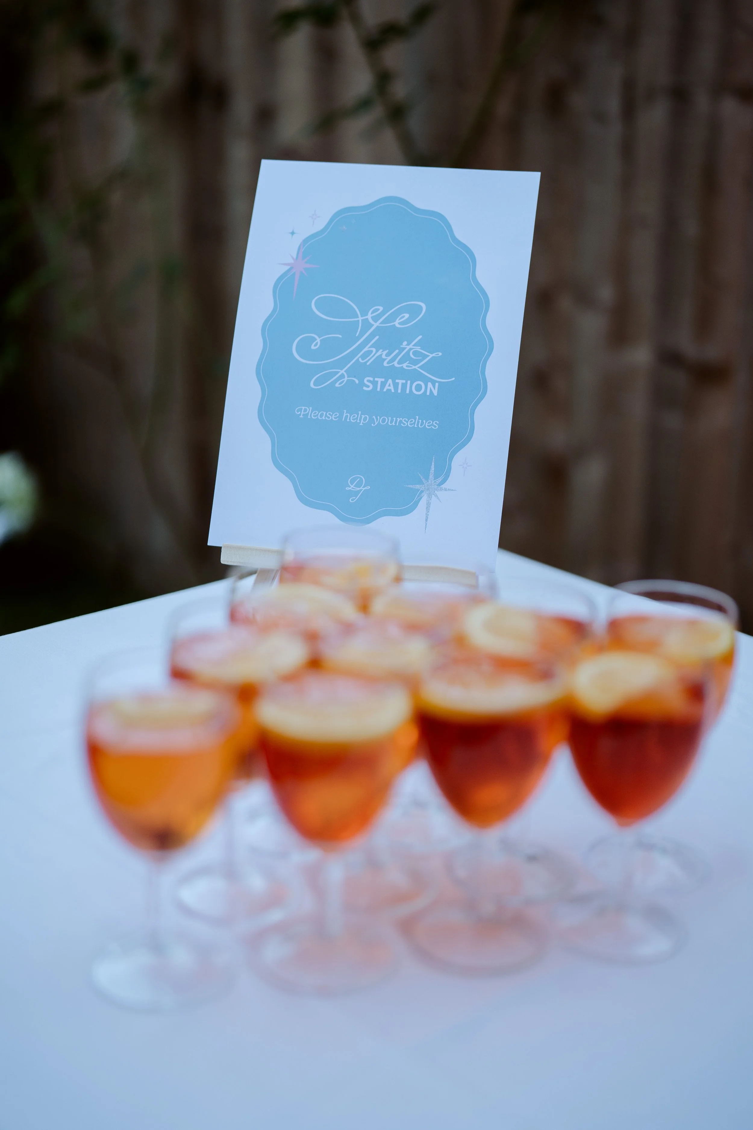A sign labeled 'Spritz Station' with a request to 'Please help yourselves' and a display of small glasses of a pinkish-orange drink on a covered table. The sign has a decorative light blue border and is set against a blurred background with a wooden 