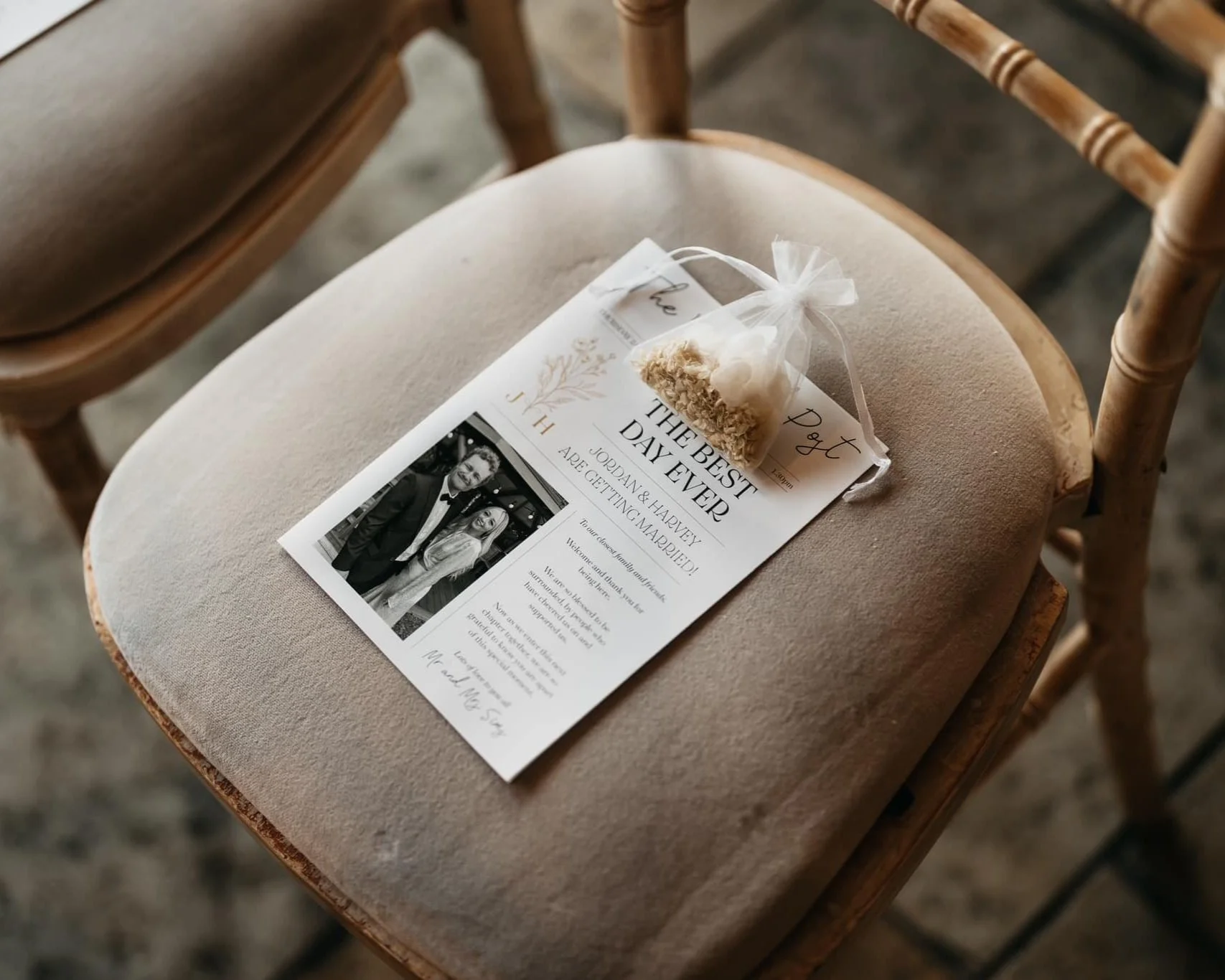 A wedding invitation on a beige cushioned chair with a small organza bag containing dried flowers placed on top of it.