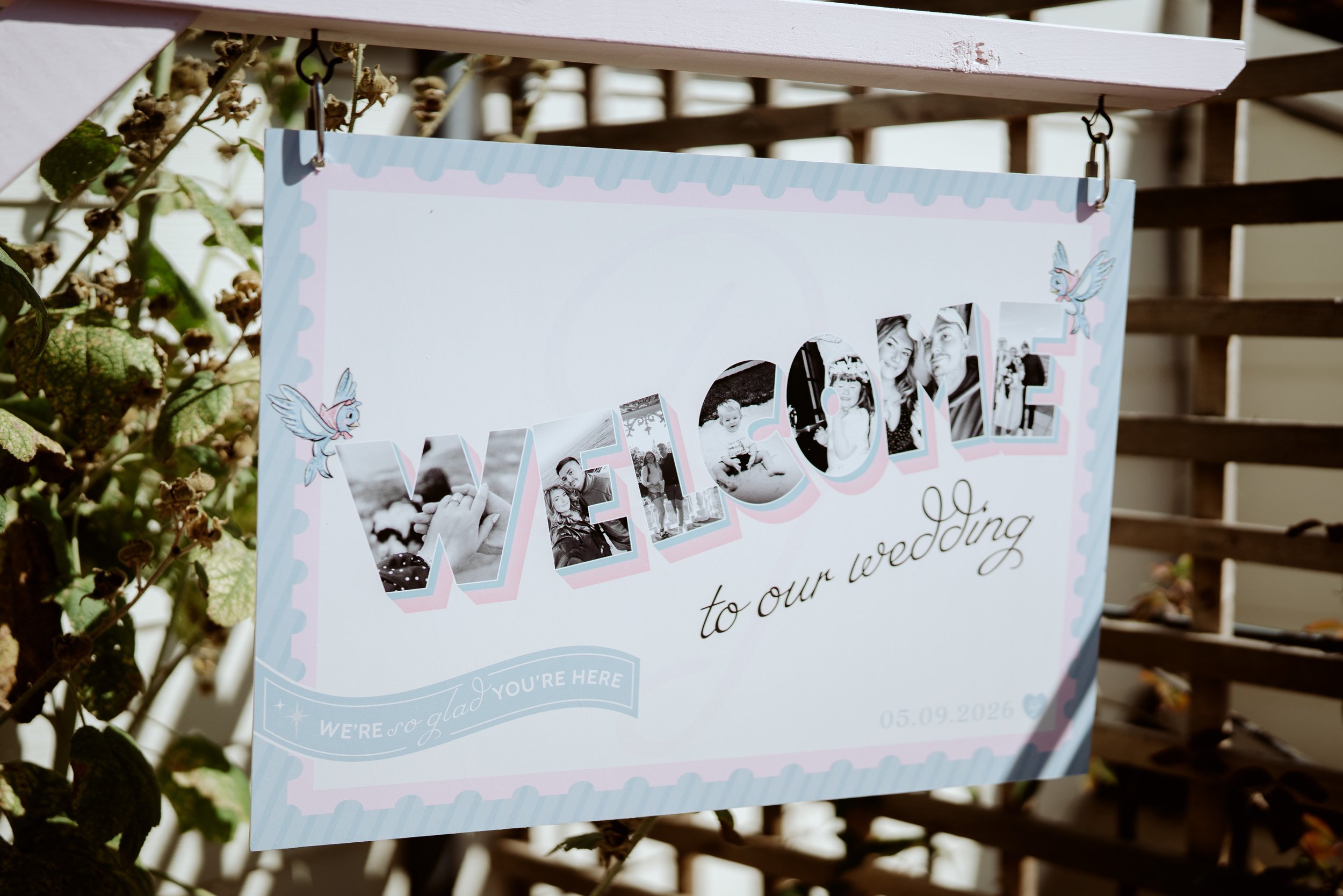 A wedding photo display with the word 'LOVE' in cut-out letters filled with black-and-white photos of a couple, decorated with fairy illustrations, hanging outdoors on a lattice fence among green leaves.