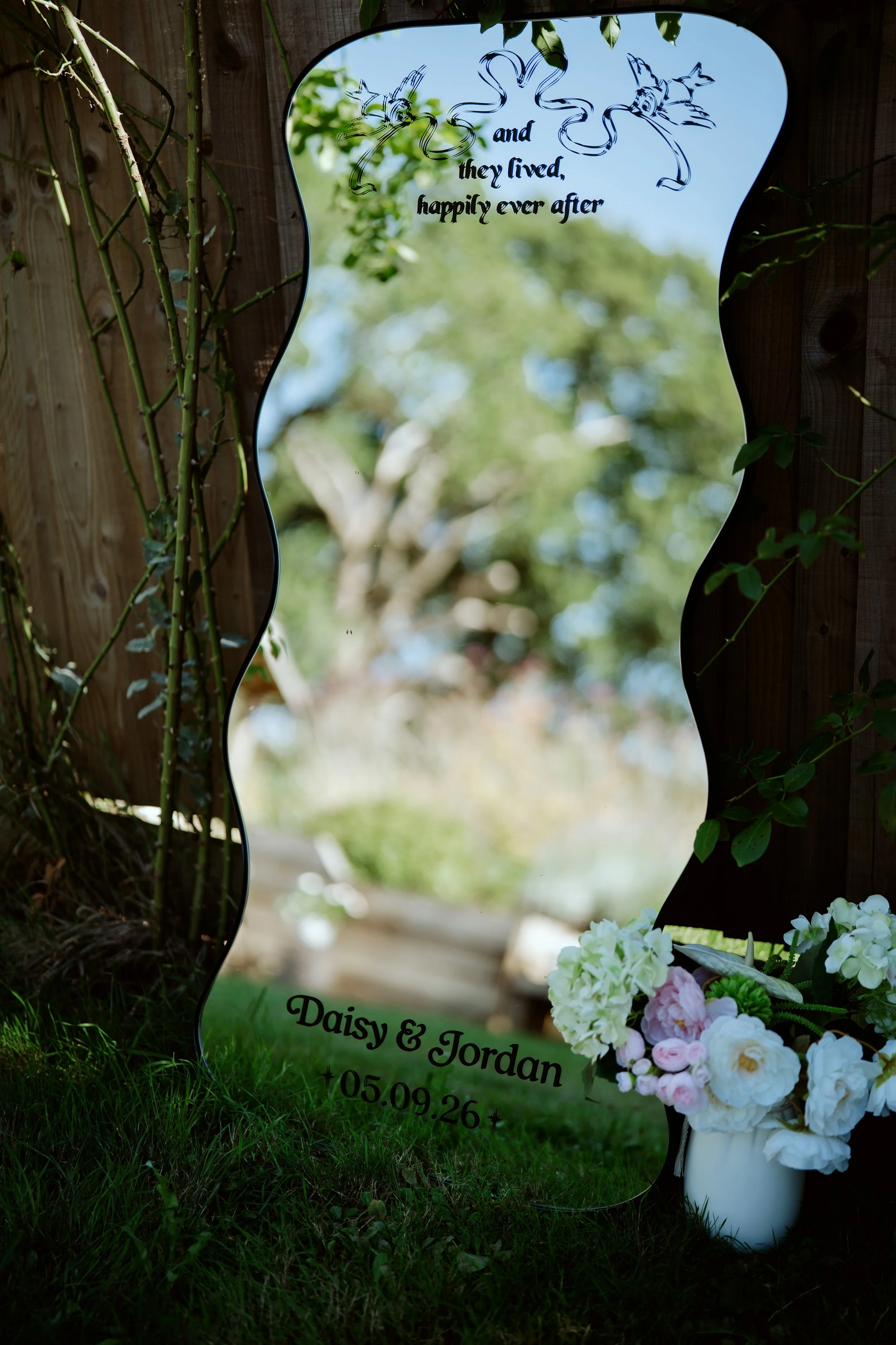 Wedding memorial sign with floral decorations and a mirror reflecting trees in the background, indicating a ceremony for Daisy and Jordan on September 26.