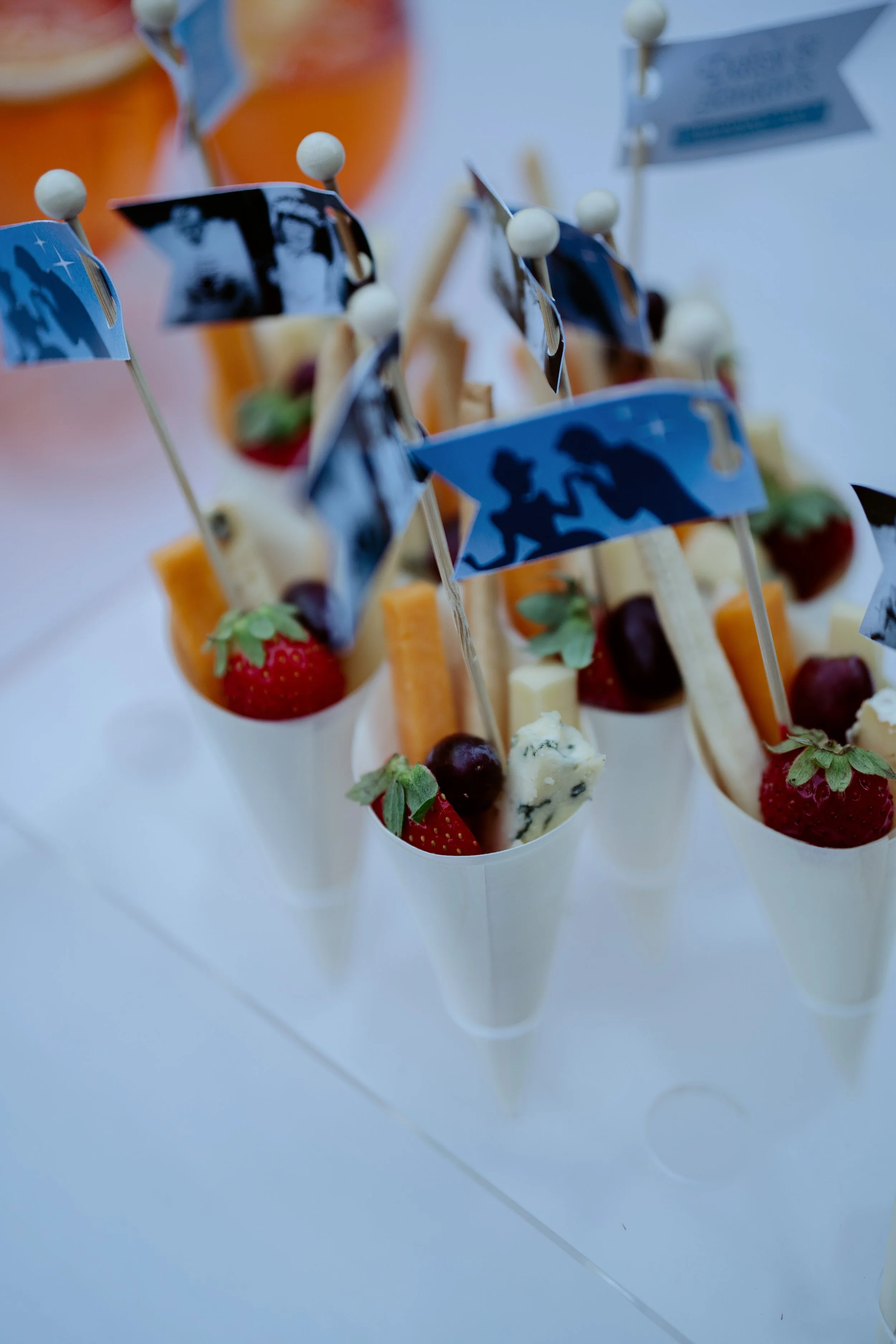 Small dessert cups filled with strawberries, blueberries, cheese, and garnished with small flags on toothpicks, arranged on a white surface.