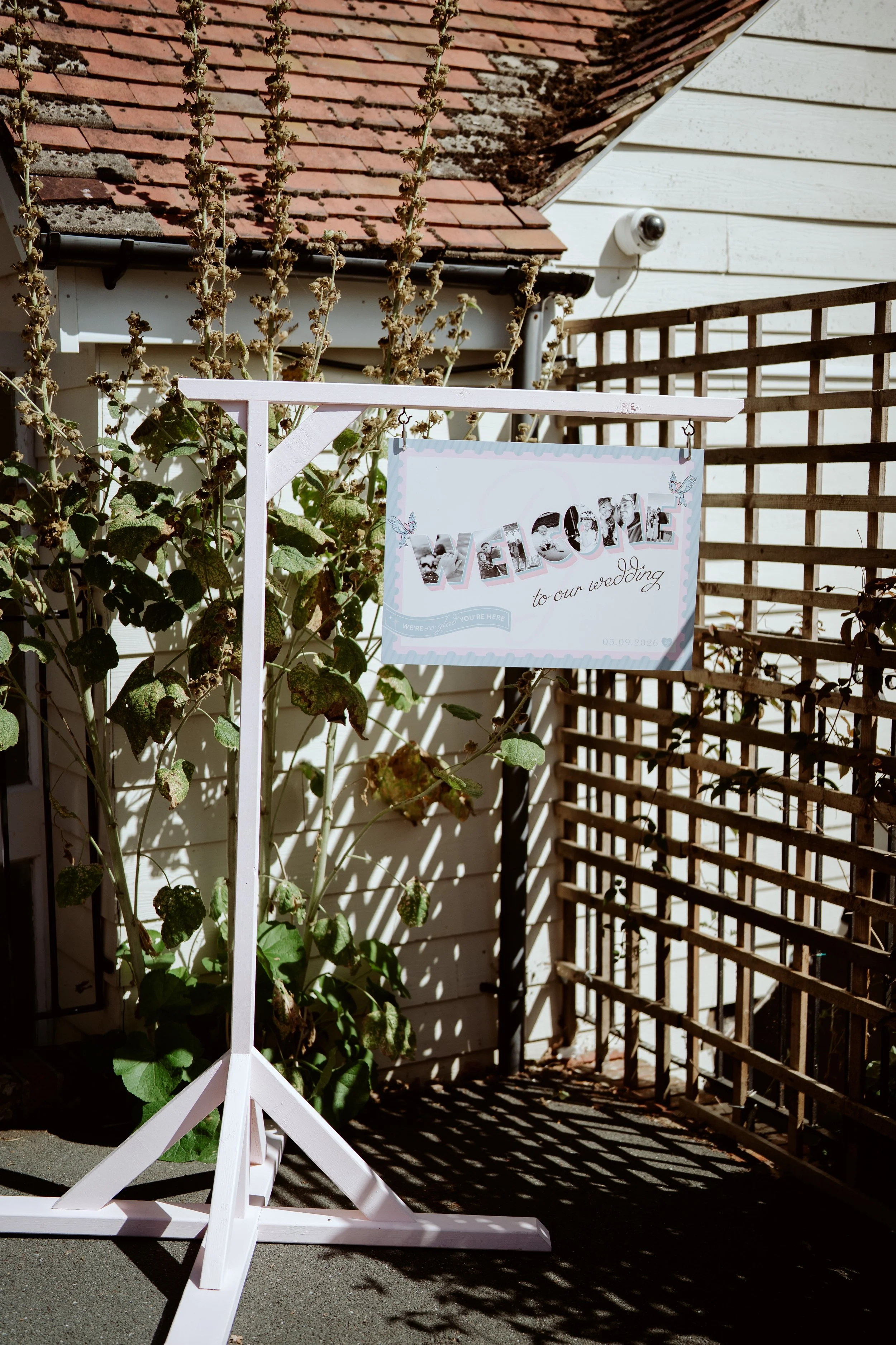 A white wooden wedding sign stand with a sign that says "Welcome to our wedding" and includes a row of black-and-white photos of a couple, set outdoors near a building with a wooden lattice fence and a white wall. Shadows of the stand and plants are 