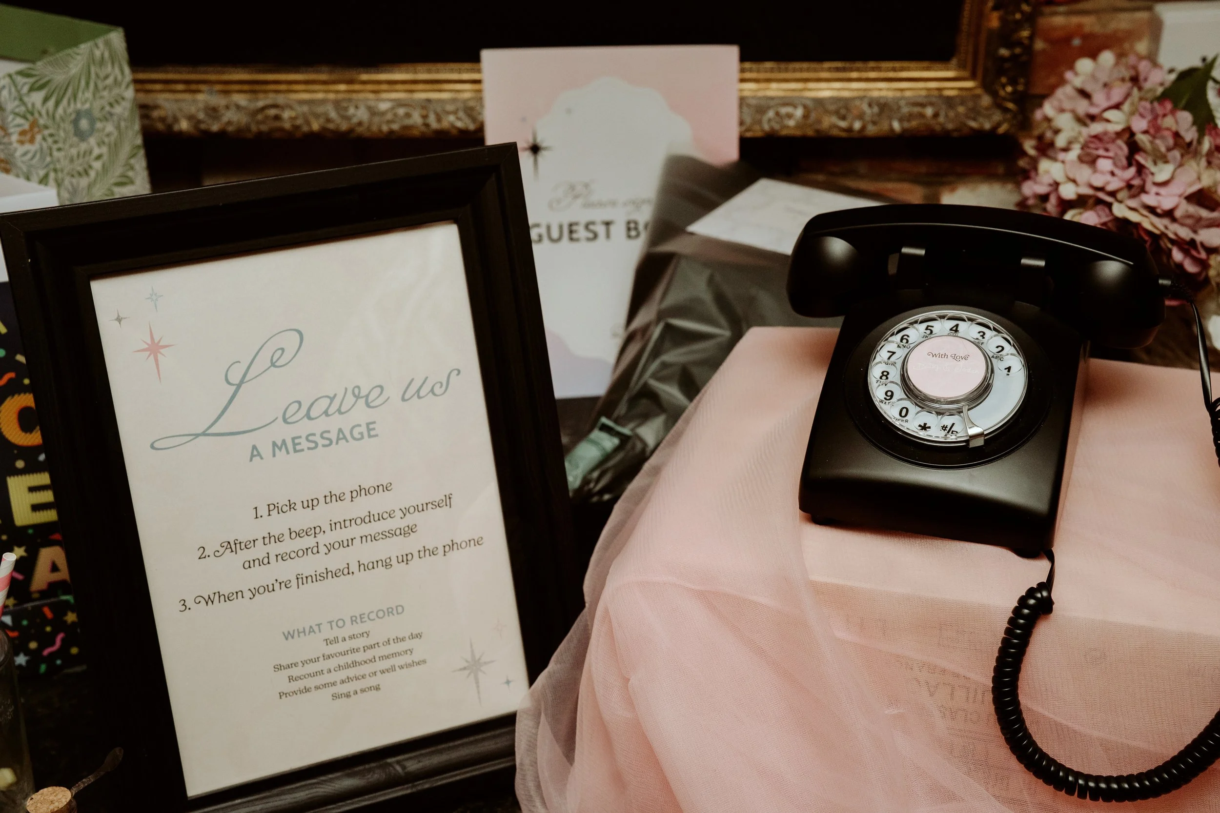 A display table with framed signs, a vintage rotary phone, and floral decorations. One sign titled "Leave Us a Message" with instructions for recording a message after a guest stays.