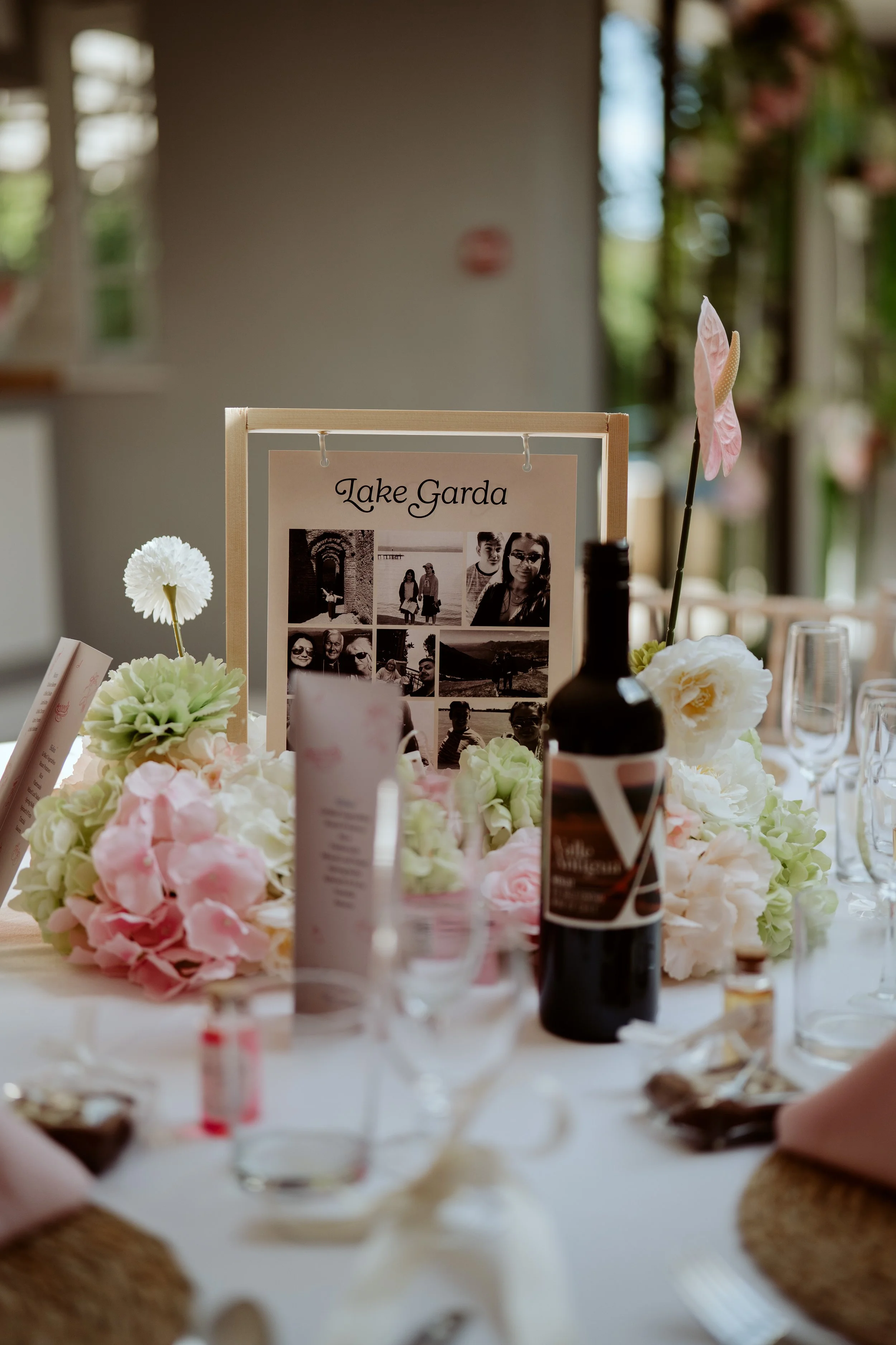A table decorated with pink and white flowers, a framed collage of black and white photos labeled 'Lake Garda,' a bottle, and various small objects, with a blurred background of windows and plants.