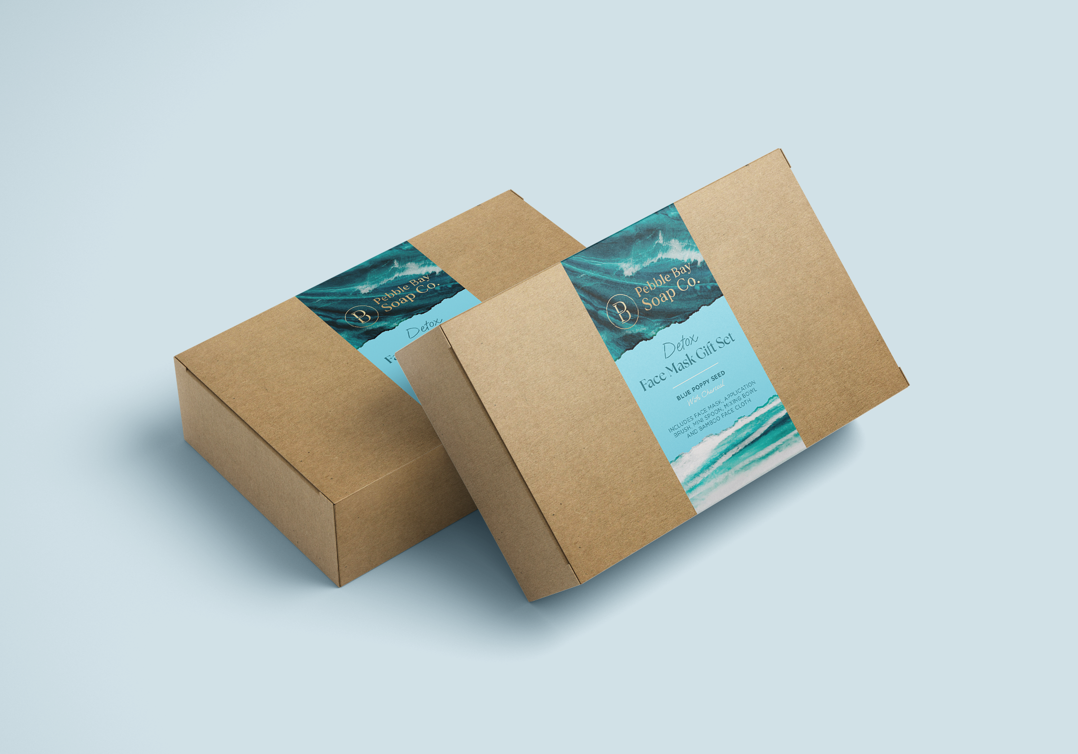 Two brown cardboard boxes labeled Peacock Bay Soap Co. Detox Face Mask Gift Set with blue poppy seed, blue ocean scene on the labels, placed on a light blue surface.