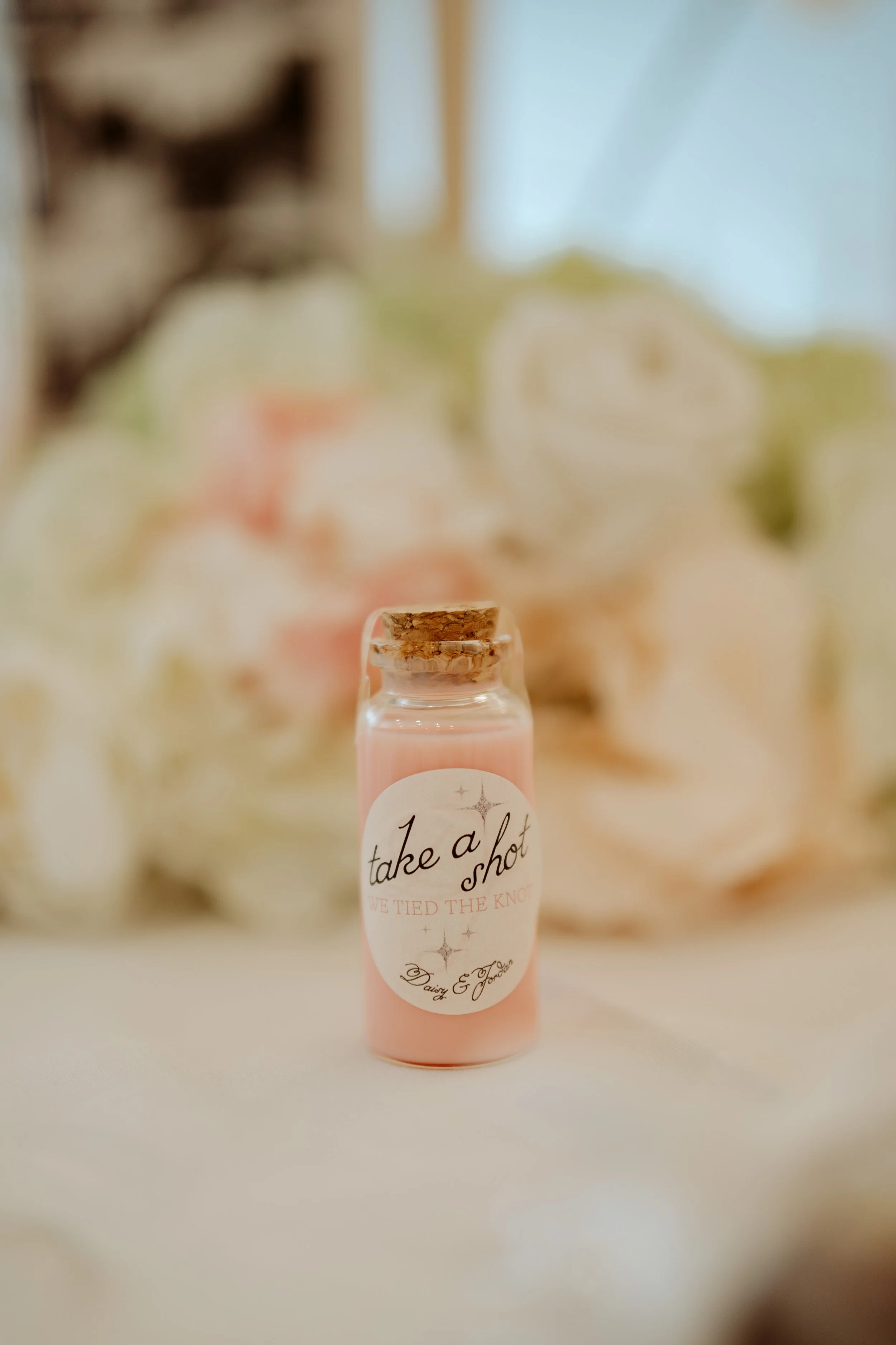Small pink bottle with a cork stopper. The label reads 'take a shot' and 'we tied the knot' with signatures below. Blurred background with floral decorations.