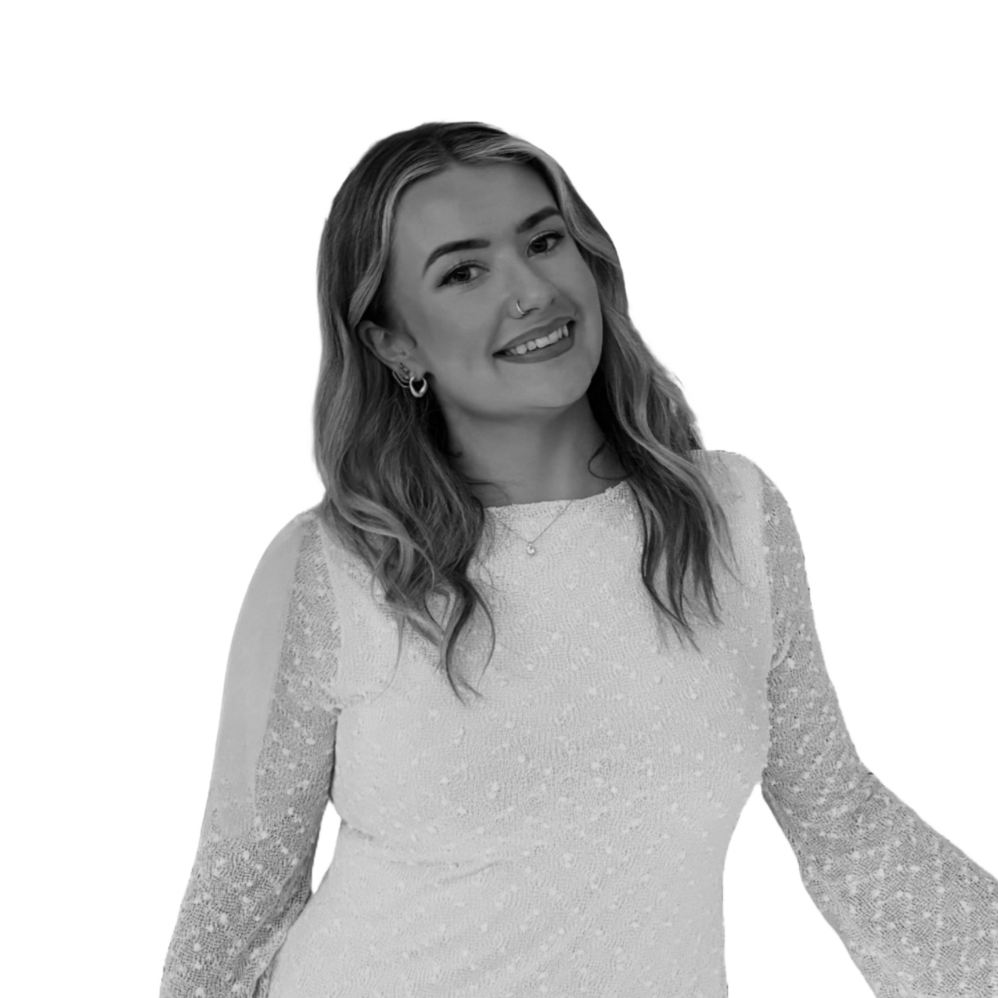 A smiling woman with long wavy hair, wearing a lace dress, hoop earrings, a nose ring, a necklace with a small pendant, and a bracelet, standing against a black background.