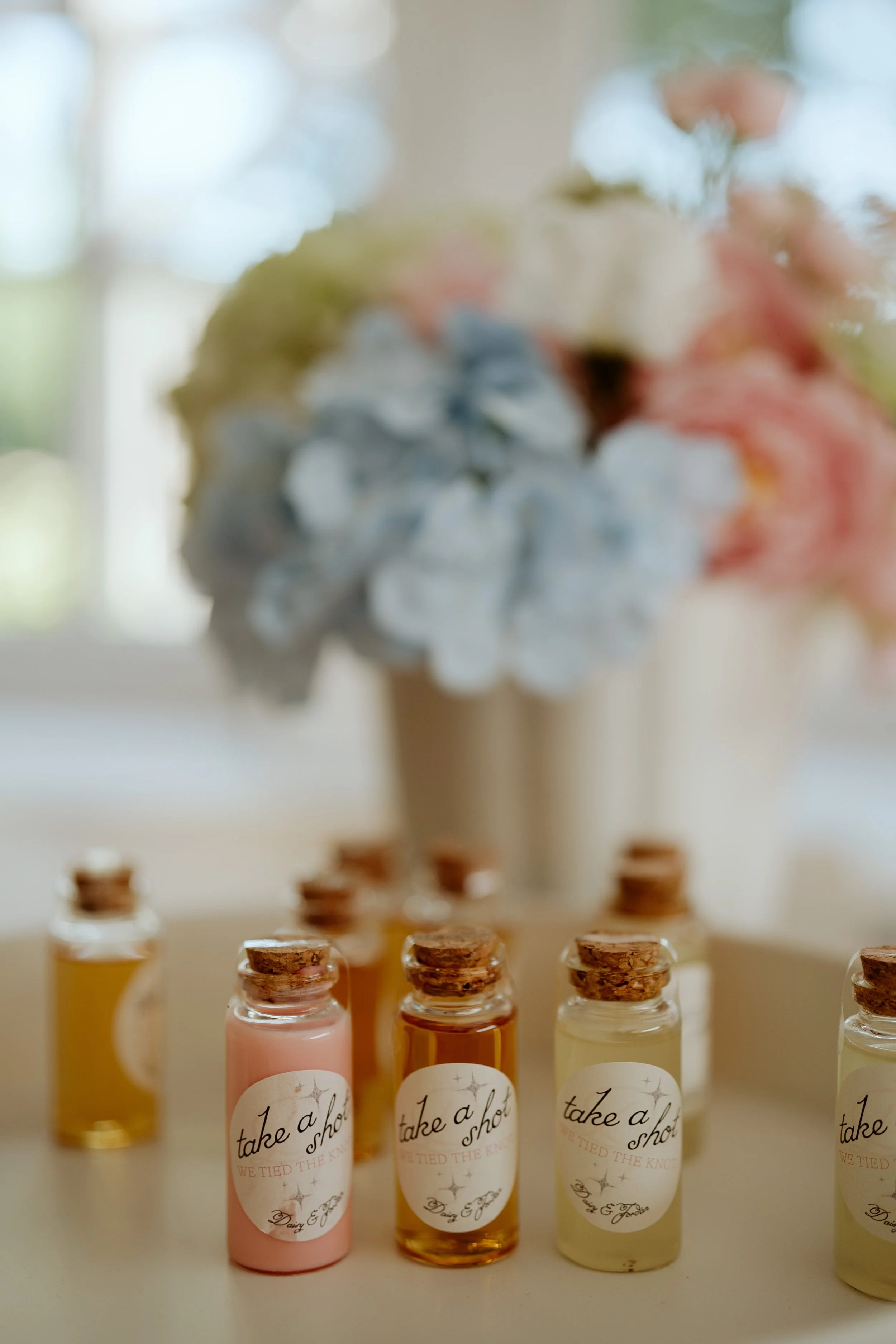 Multiple small glass bottles with cork stoppers arranged on a surface, with a blurred floral arrangement in the background.