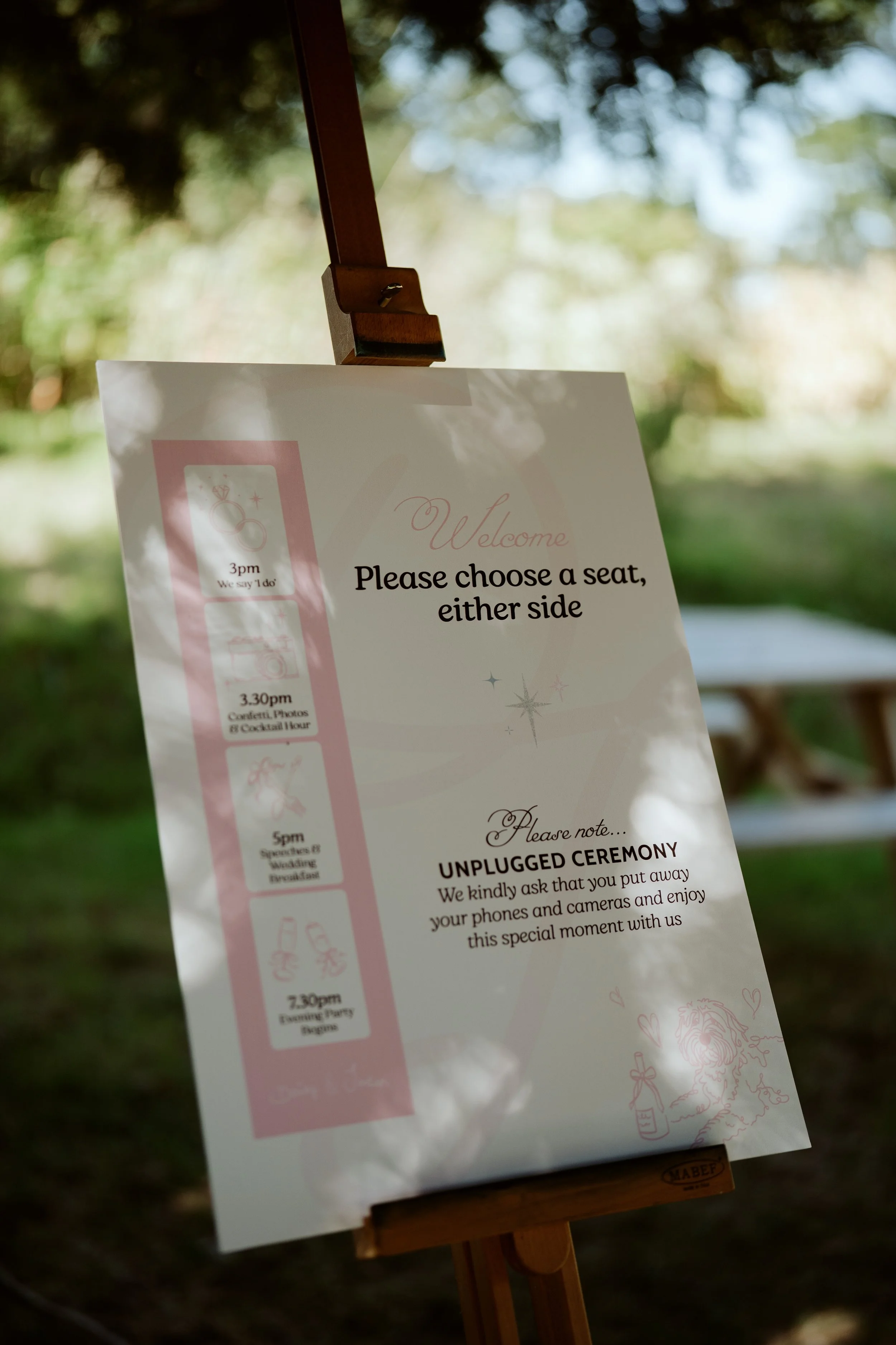 A sign with wedding ceremony details, including a schedule and reminders to unplug electronic devices, hangs on a wooden stand outdoors with blurred trees and sunlight in the background.