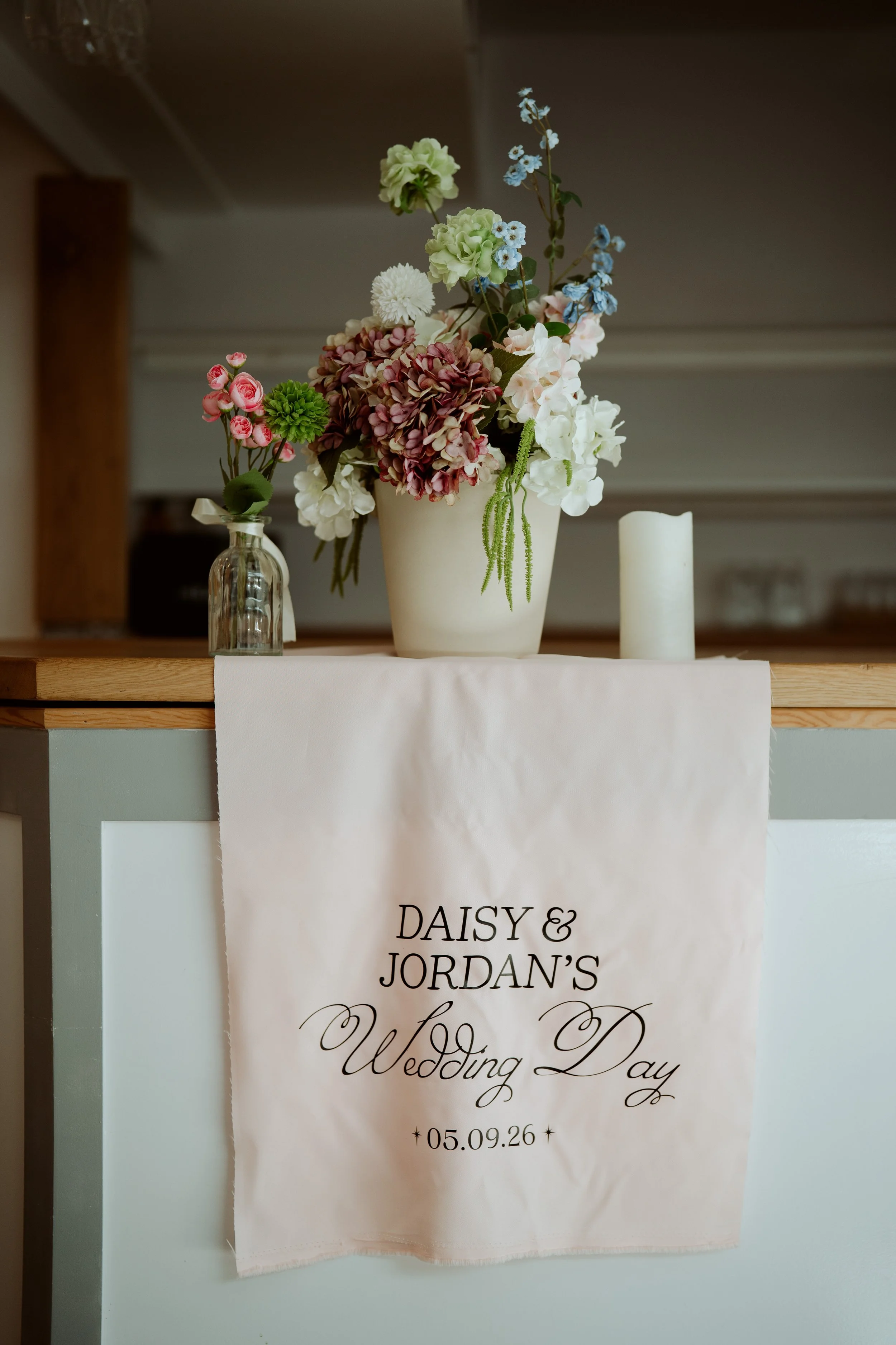 A white table with a flower arrangement in a white vase, a small clear glass bottle with pink flowers, and a white candle. A fabric banner with elegant black script that reads "Daisy & Jordan's Wedding Day" and the date "05.09.26" hangs on the front 