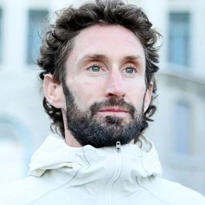 Man with curly hair and beard wearing a white jacket outdoors.