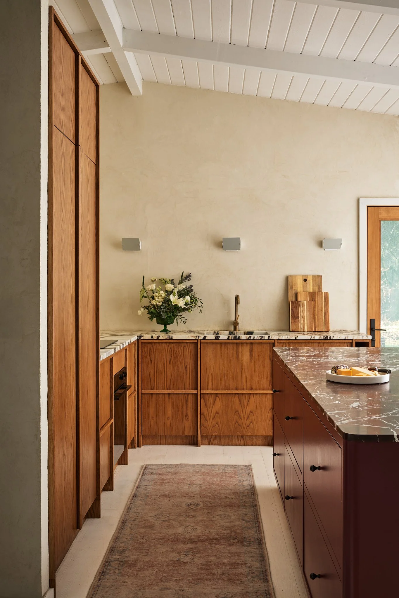 A cozy kitchen with wooden cabinets, a central island with a dark marble countertop, a bouquet of white flowers on the counter, and a doorway with a window showing a garden or green outdoor scenery.