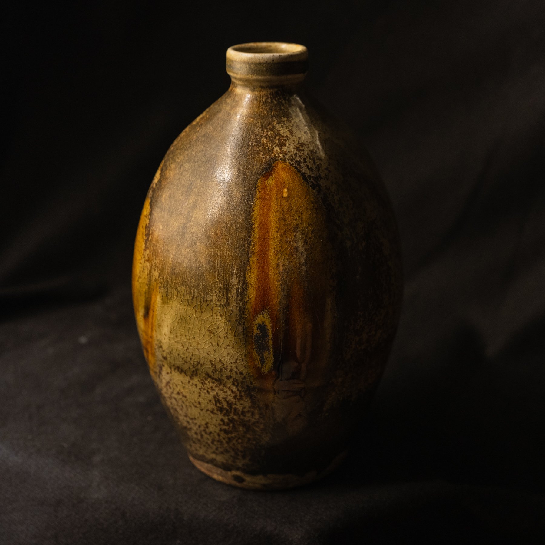 A rustic, handmade ceramic vase with a rounded body and narrow neck, featuring earth-tone glazes and crackle patterns, set against a black background.