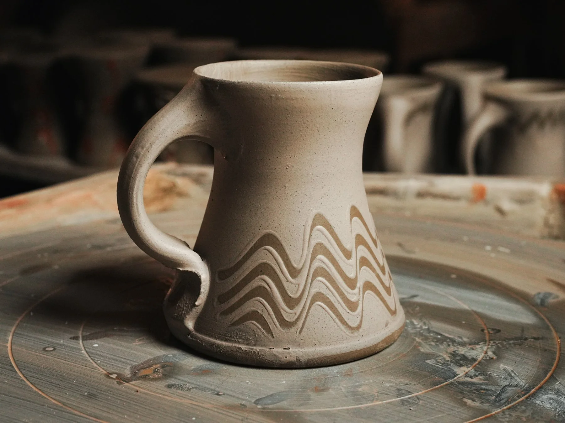 A ceramic mug with a wavy pattern along its body, placed on a pottery wheel in a pottery studio.