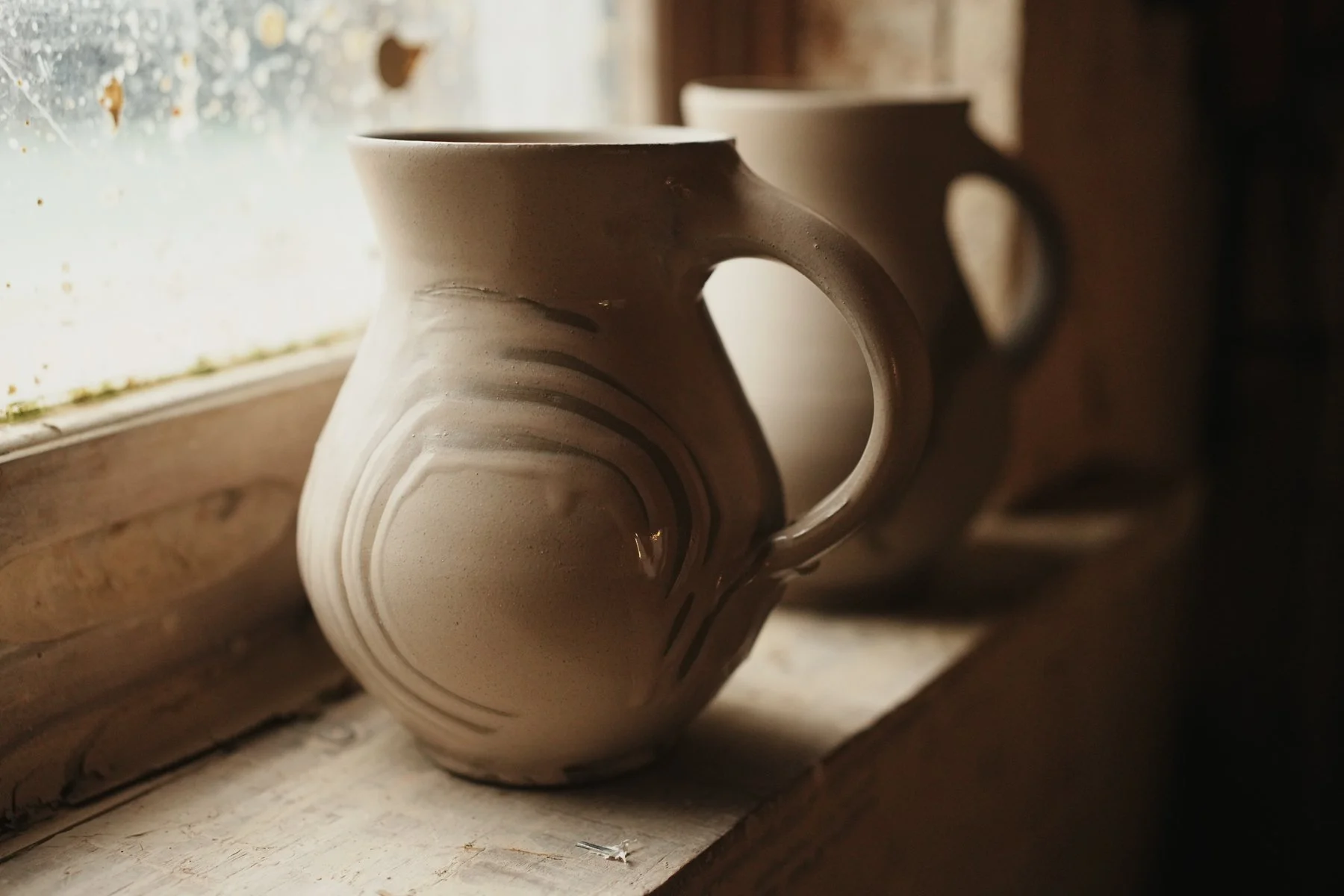 A beige ceramic mug with black curved lines, sitting on a wooden window sill with a frosted window behind it, in warm natural lighting.