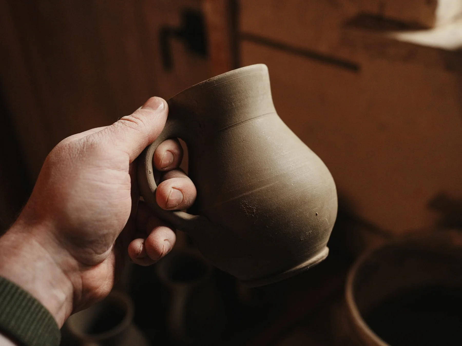 Close-up of a person's hand holding a small, unfinished ceramic mug in a pottery studio with other clay mugs in the background.