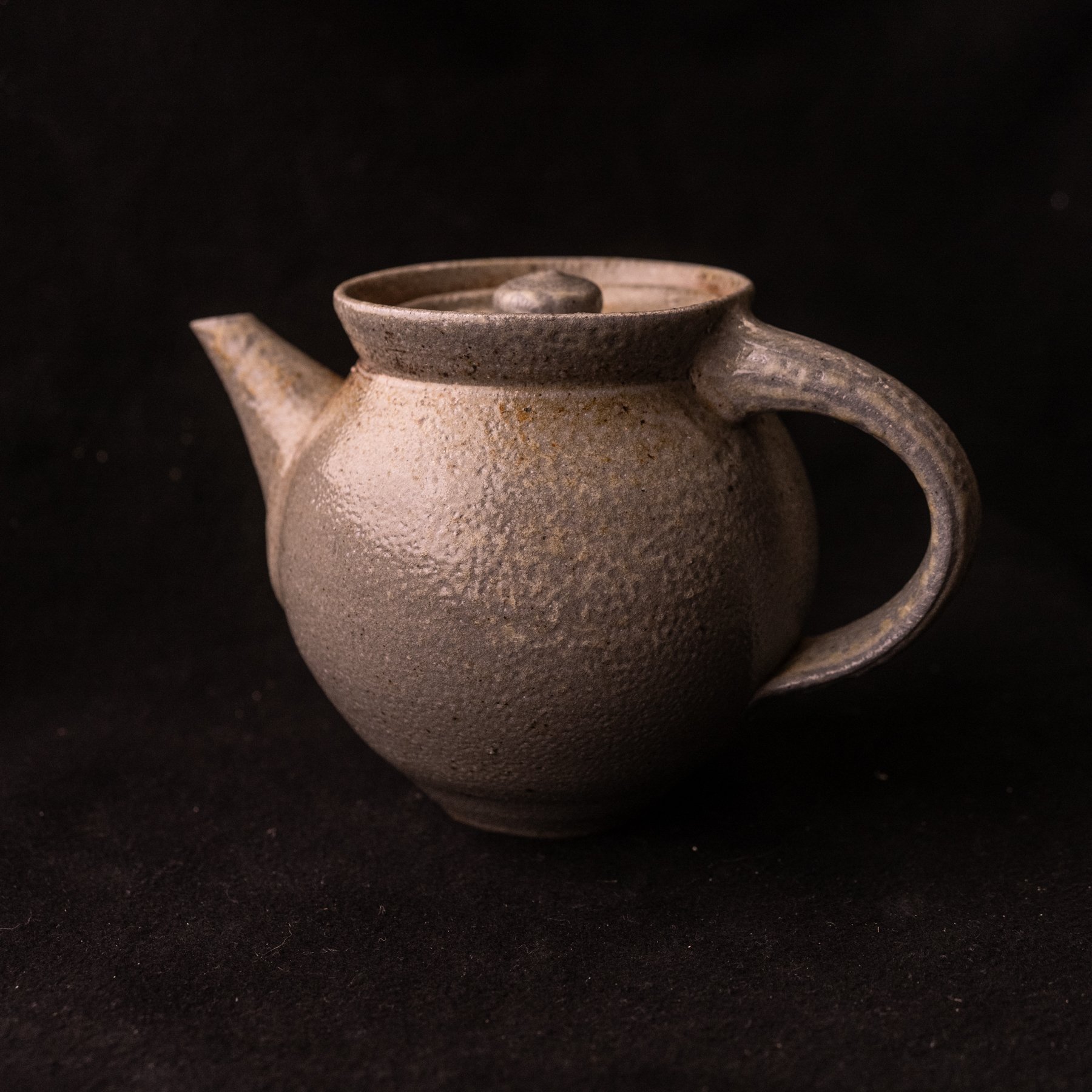 A rustic ceramic teapot with a rounded body, a short spout, a curved handle, and a lid with a knob, set against a black background.