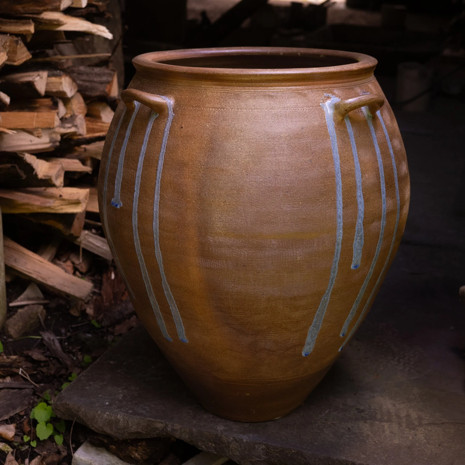 A large ceramic vase with vertical blue lines on a tan surface, placed outdoors next to a stack of firewood on a stone ledge.