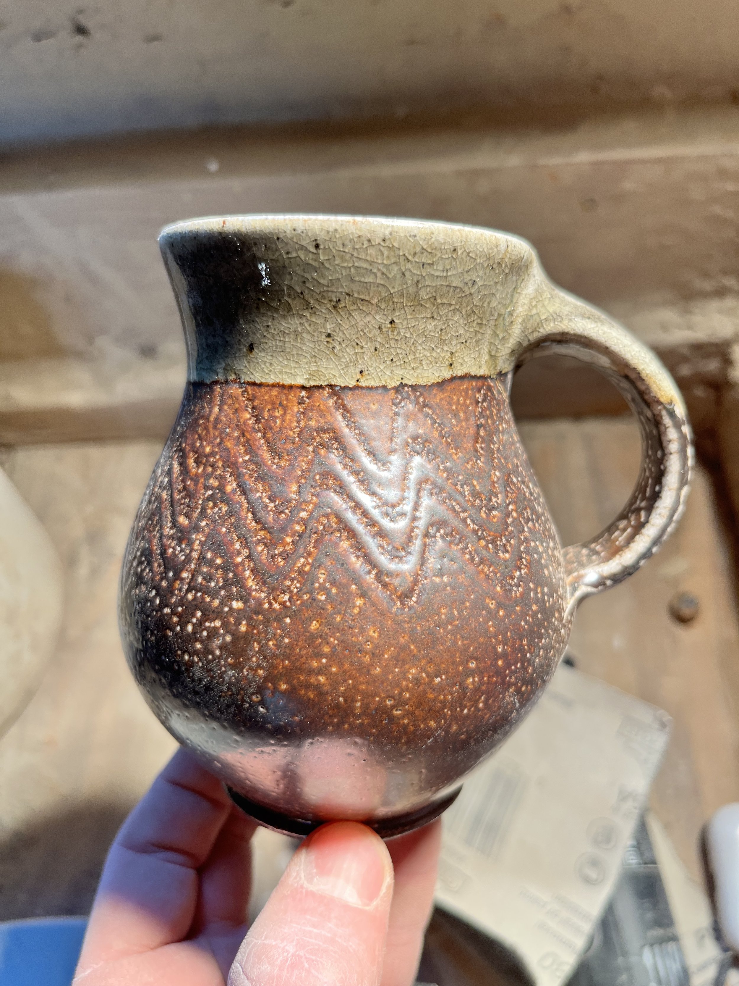 Close-up of a ceramic mug with a textured, reddish-brown pattern and a dark glaze on the rim, being held by a person's hand.
