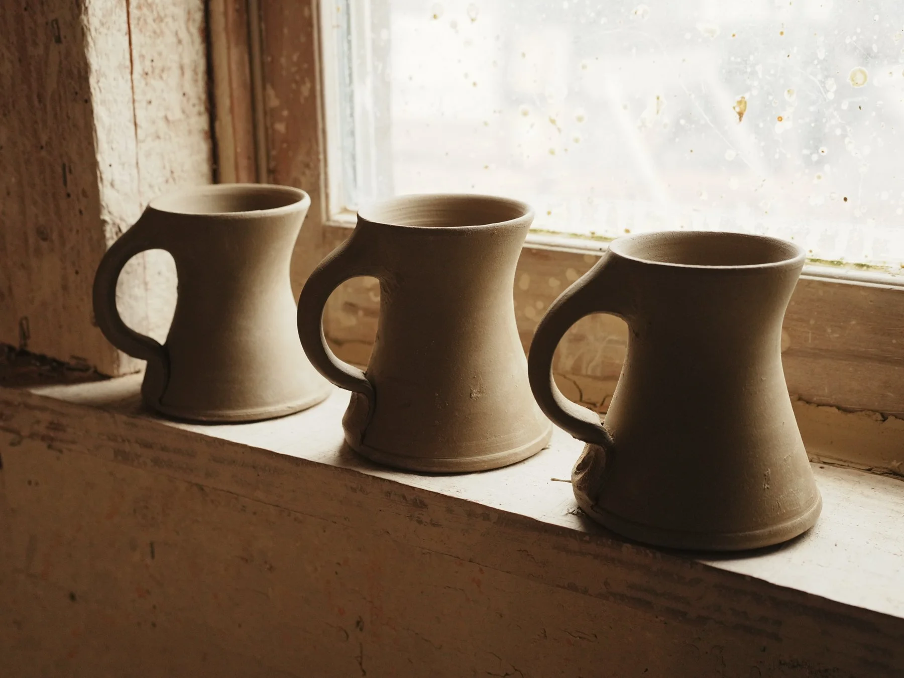 Three unfinished ceramic mugs with handles sit on a windowsill inside a rustic building. Sunlight streams through a slightly foggy window, illuminating the mugs and creating soft shadows.