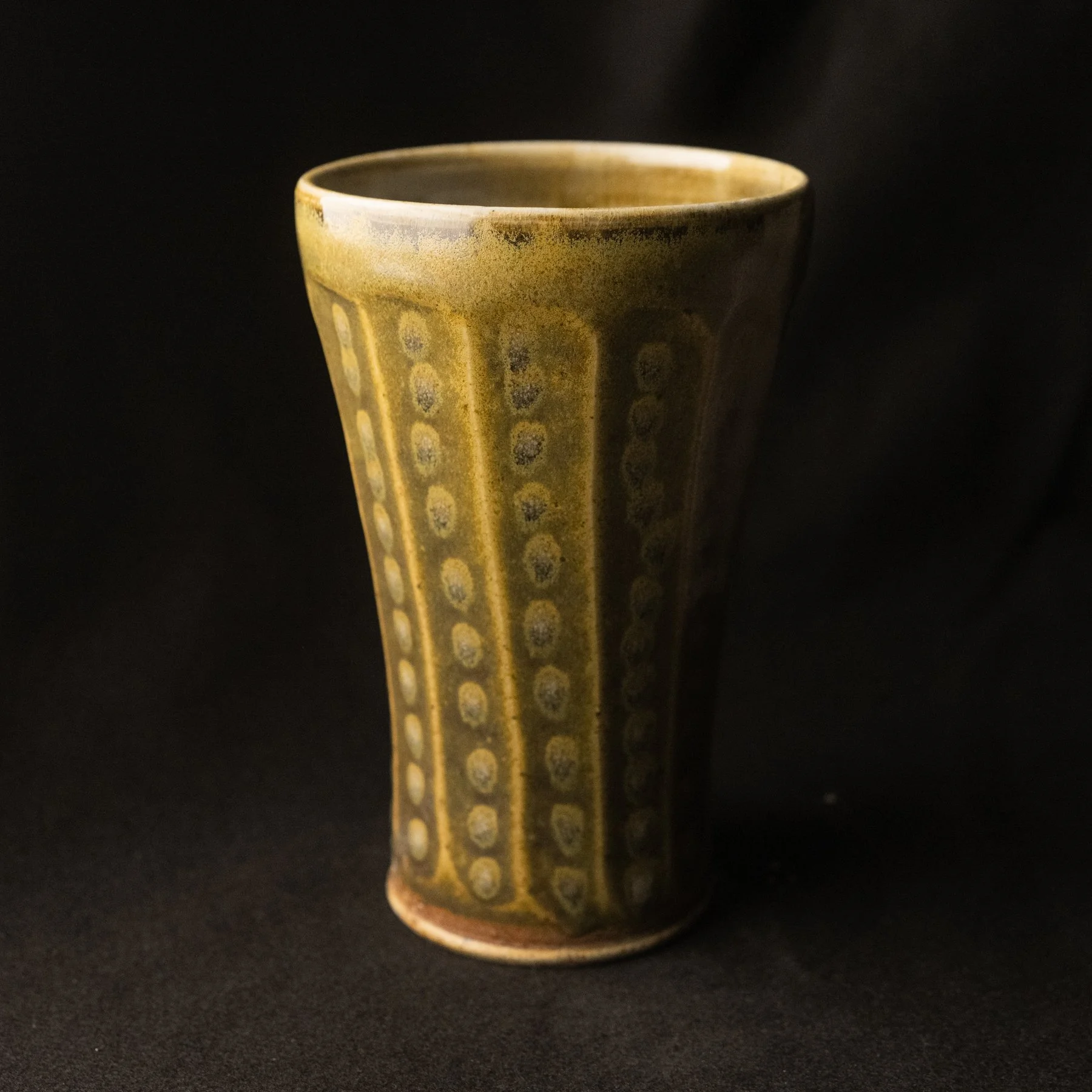 A ceramic cup with a yellowish glaze and dotted pattern, set against a black background.