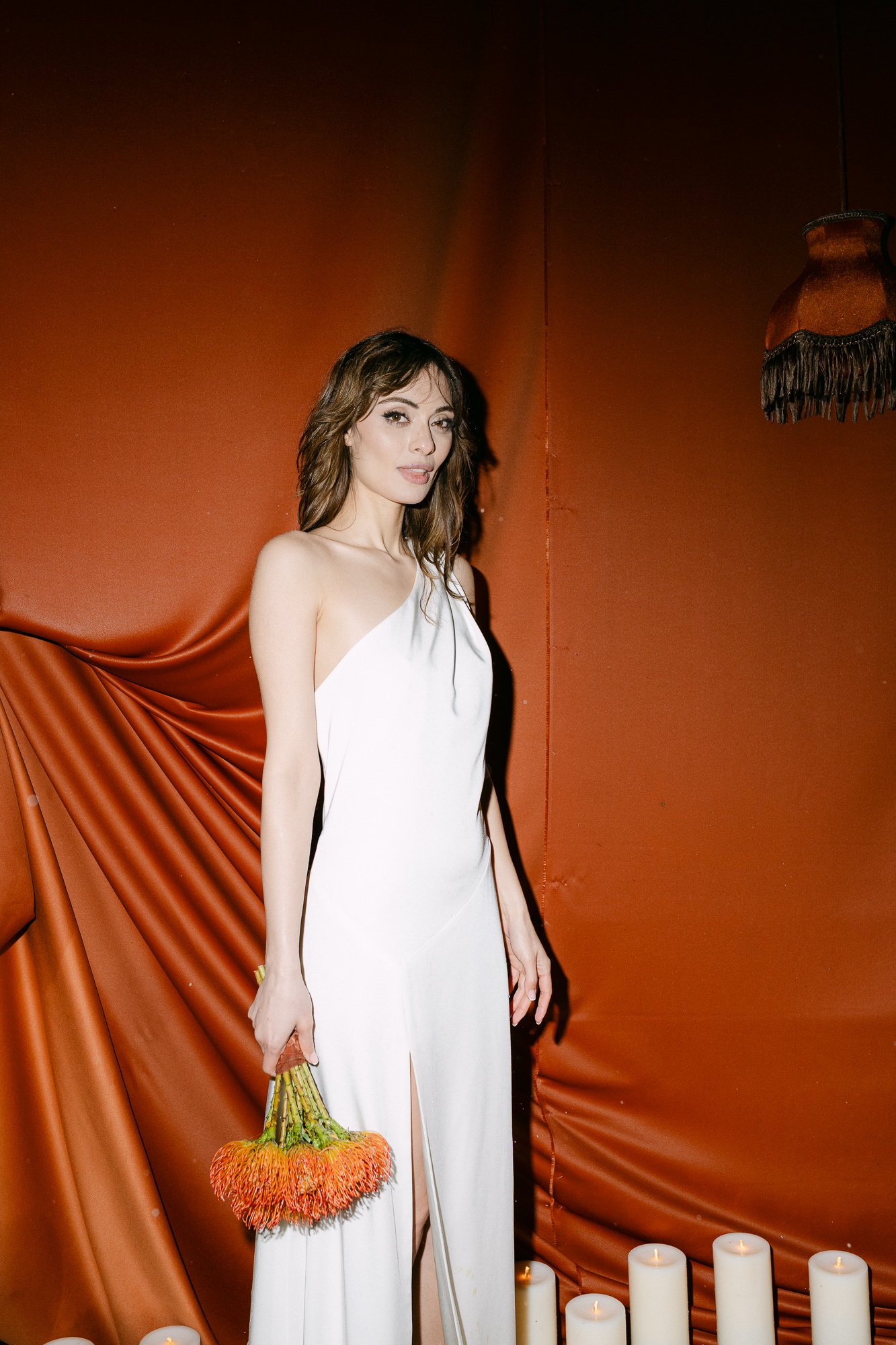 Bride in white dress standing in front of deep orange wall holding yellow flowers — Tuscany