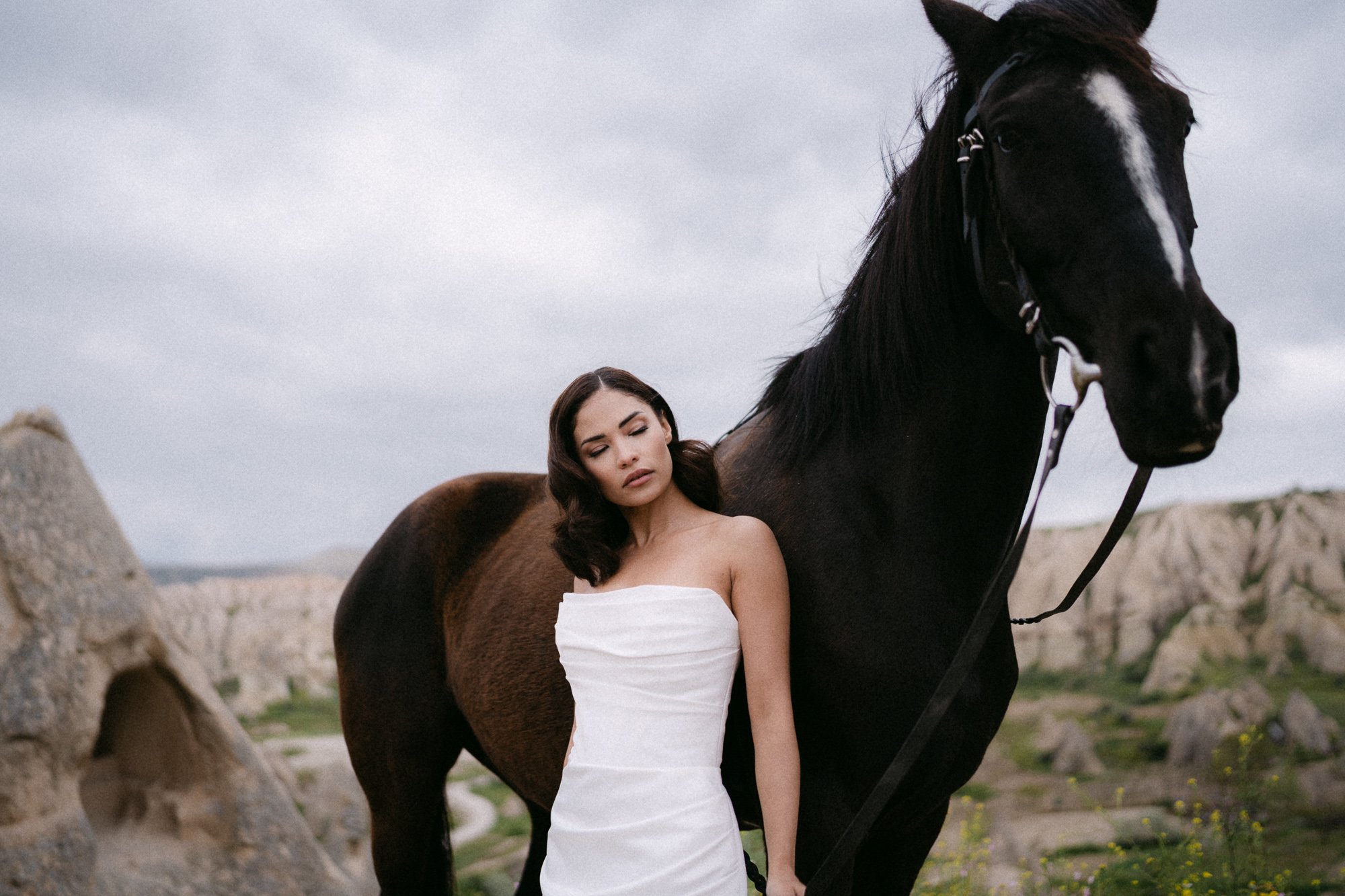 Bride in white dress standing beside black horse in Cappadocia landscape