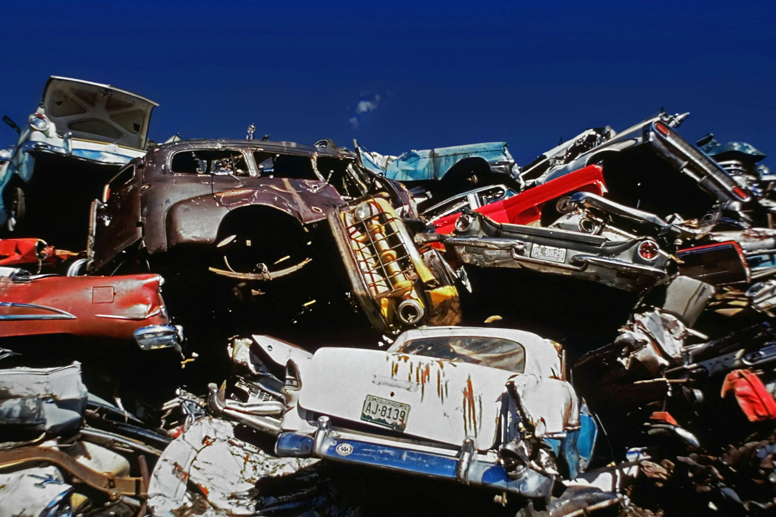 A large pile of crushed and rusted vintage cars stacked in a junkyard under a clear blue sky, showcasing various colors and classic designs from past decades.
