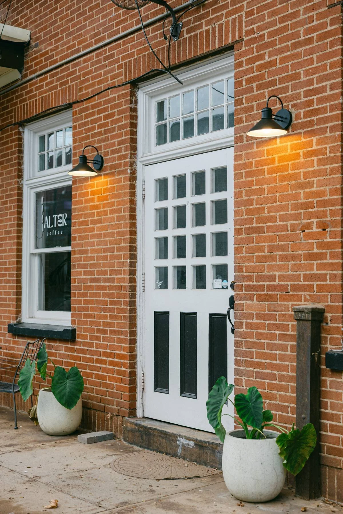 A brick building with white-framed windows and a white double door with glass panes. Two outdoor wall lights illuminate the door. There are large potted plants with green leaves on the sidewalk in front of the building, and a black metal chair is vis