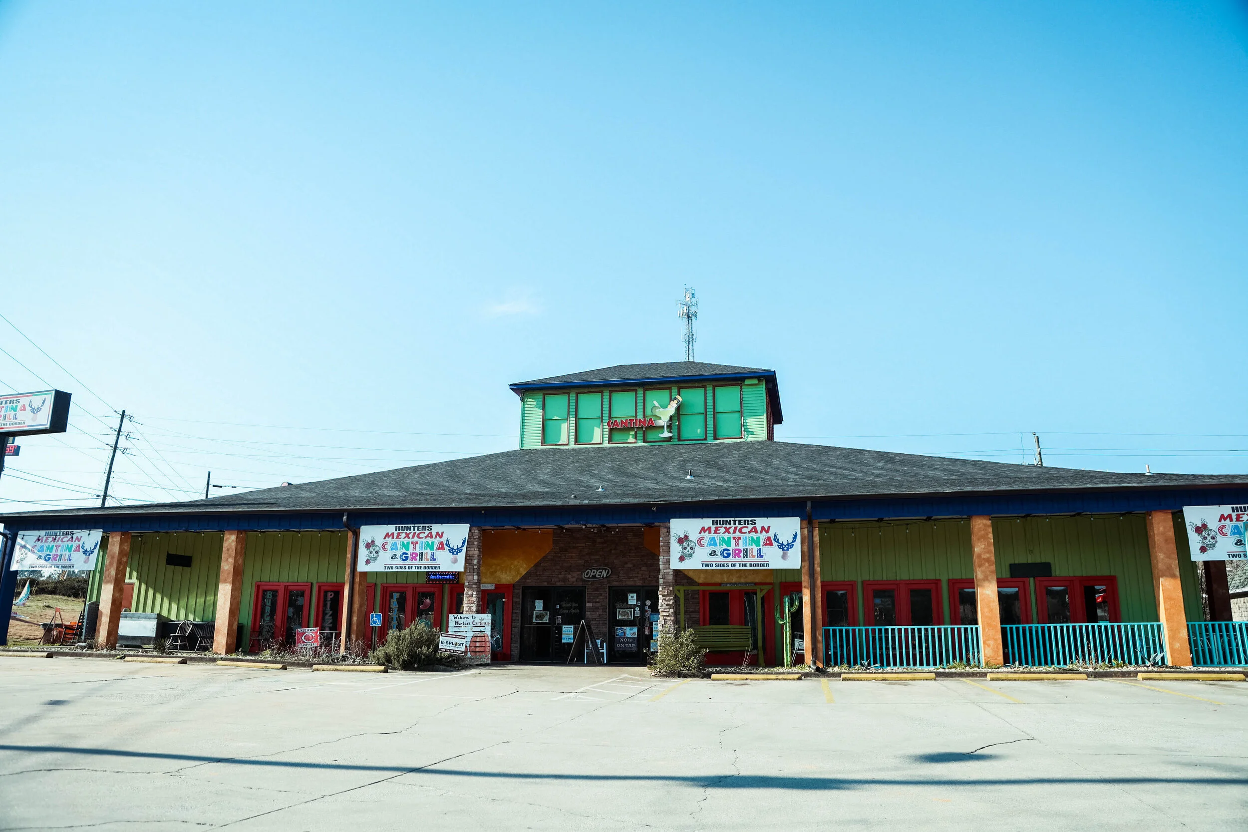 Exterior of a Mexican cantina restaurant with colorful signs and banners, parking lot in front, and a distinctive green upper facade with a rooster sculpture.
